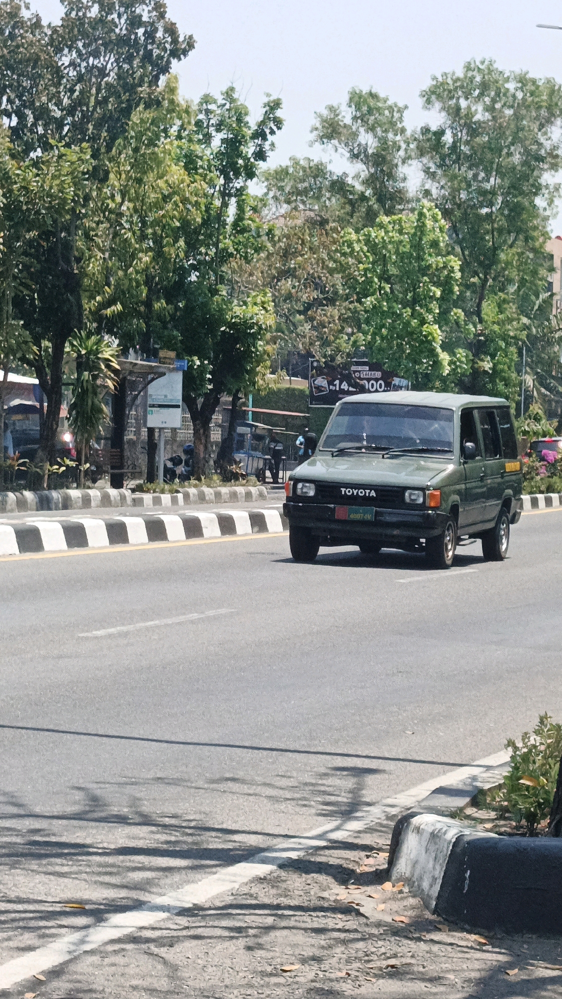 An overcast, daytime, medium shot captures a dark green Toyota Kijang van driving on a paved road in Surakarta, Indonesia. The van is facing forward, slightly angled to the right, with its headlights on. The van has a visible "TOYOTA" logo on the front grill and a license plate that reads "B 8827 IV". The van is on the right lane of a two-lane road. The road is bordered by a sidewalk with black and white striped dividers. To the left of the road, there are several trees with lush green foliage, and a bus stop shelter. In the background, there are more trees and a few buildings. The sky is a pale blue with some scattered clouds. The overall impression is a typical urban street scene in Indonesia.