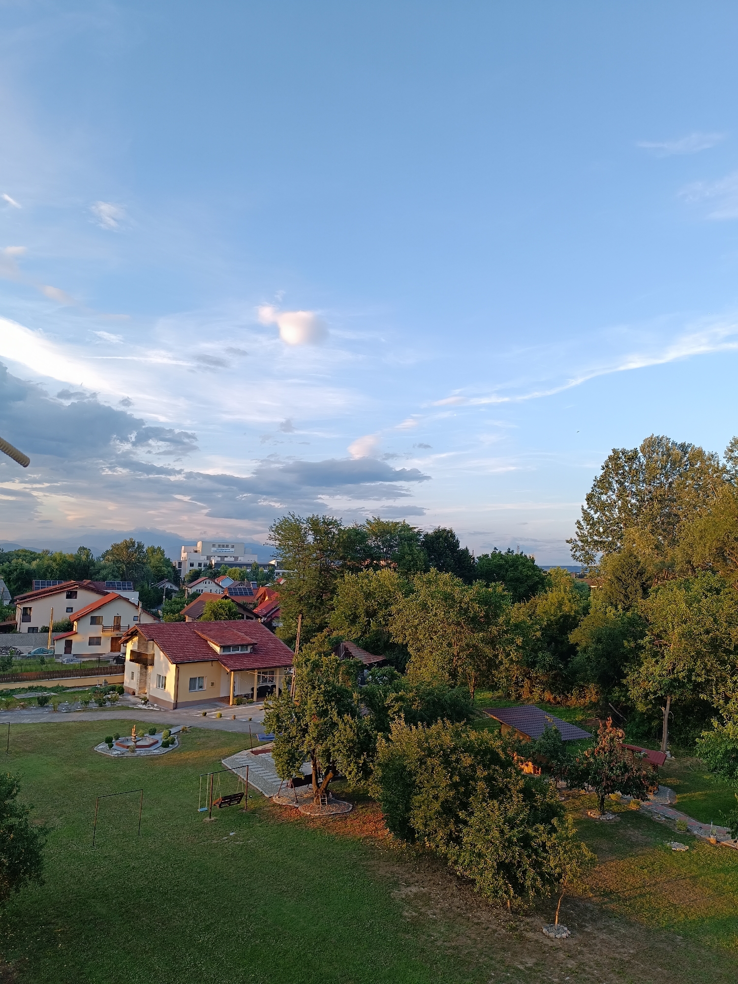 The image shows an aerial view of a residential area with houses and trees, likely in Târgu Jiu, Romania. The scene is bathed in the warm light of either sunrise or sunset, casting long shadows across the green grass of a yard. Several houses with red tiled roofs are visible, some with solar panels on their rooftops. A large, modern white building with multiple windows stands in the background. The yard in the foreground features a fountain, two swings, and a paved path leading to a small structure with a brown roof. Lush green trees of various sizes surround the houses and the yard. The sky is a pale blue with scattered white clouds, some of which are illuminated by the sun. There are no people or animals visible in the image.