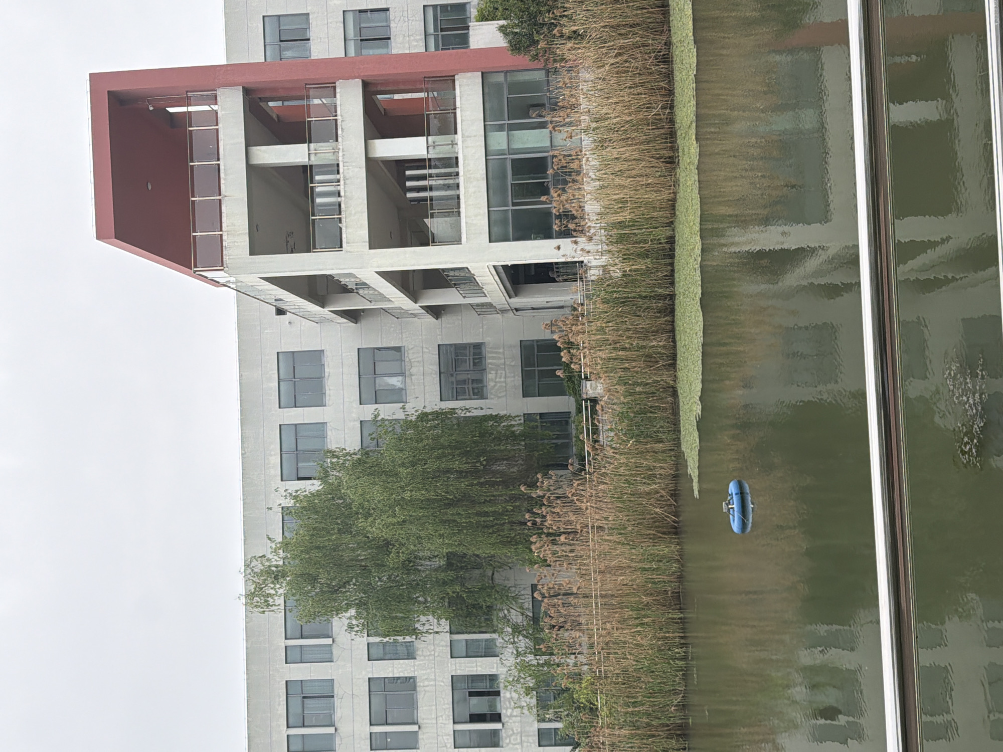 The image shows a large, modern building with a red and white exterior, likely located in the Jiangning District of China. The building has multiple floors and numerous windows. A body of water, possibly a river or a pond, is located adjacent to the building, lined with tall grasses. A small, blue inflatable object floats on the water's surface. The sky is overcast. The image is taken from an elevated position looking down at the scene.