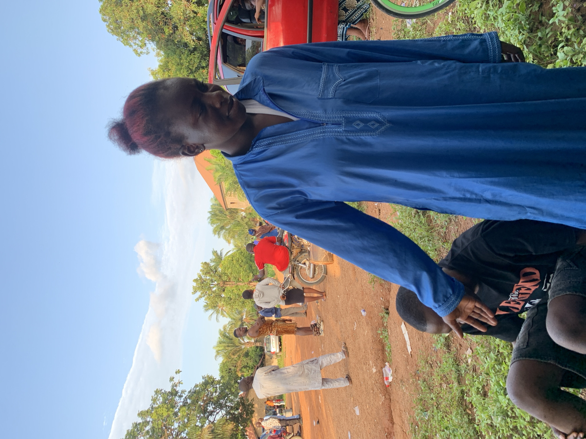 The image shows a woman in a blue tunic standing in what appears to be an outdoor setting. She has dark hair with red highlights and is facing a man who is bent over. The scene is somewhat crowded with other people in the background, some standing near motorcycles and a red car. The ground is a dirt path or road. The location is suggested to be Akwanga, Nigeria. The interaction between the woman and the bent-over man is unclear but appears to be an engagement or assistance of some kind. The environment includes trees and a partly cloudy sky.