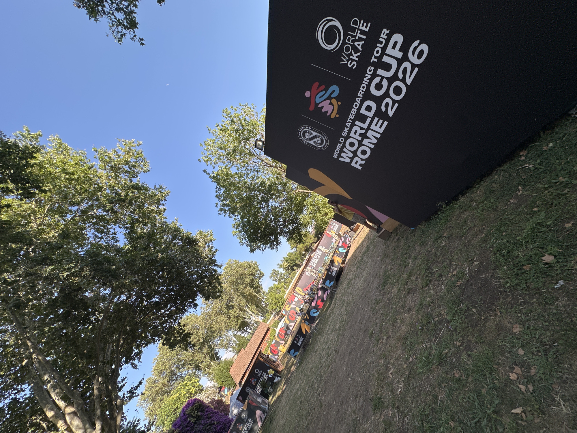 An outdoor scene with a clear blue sky above. In the foreground, a large black sign displays white text that reads "WORLD SKATEBOARDING TOUR WORLD CUP ROME 2026" along with logos. To the left of the sign, a grassy area with scattered leaves leads to a long, colorful banner decorated with various designs and text, including "WORLD CUP ROME 2026." Behind the banner, a building with a tiled roof is visible, and further back, large trees with lush green foliage dominate the landscape. Several people are present in the background, some standing near the building and others near the banner, appearing to be spectators or participants. The overall setting suggests an outdoor event or venue.