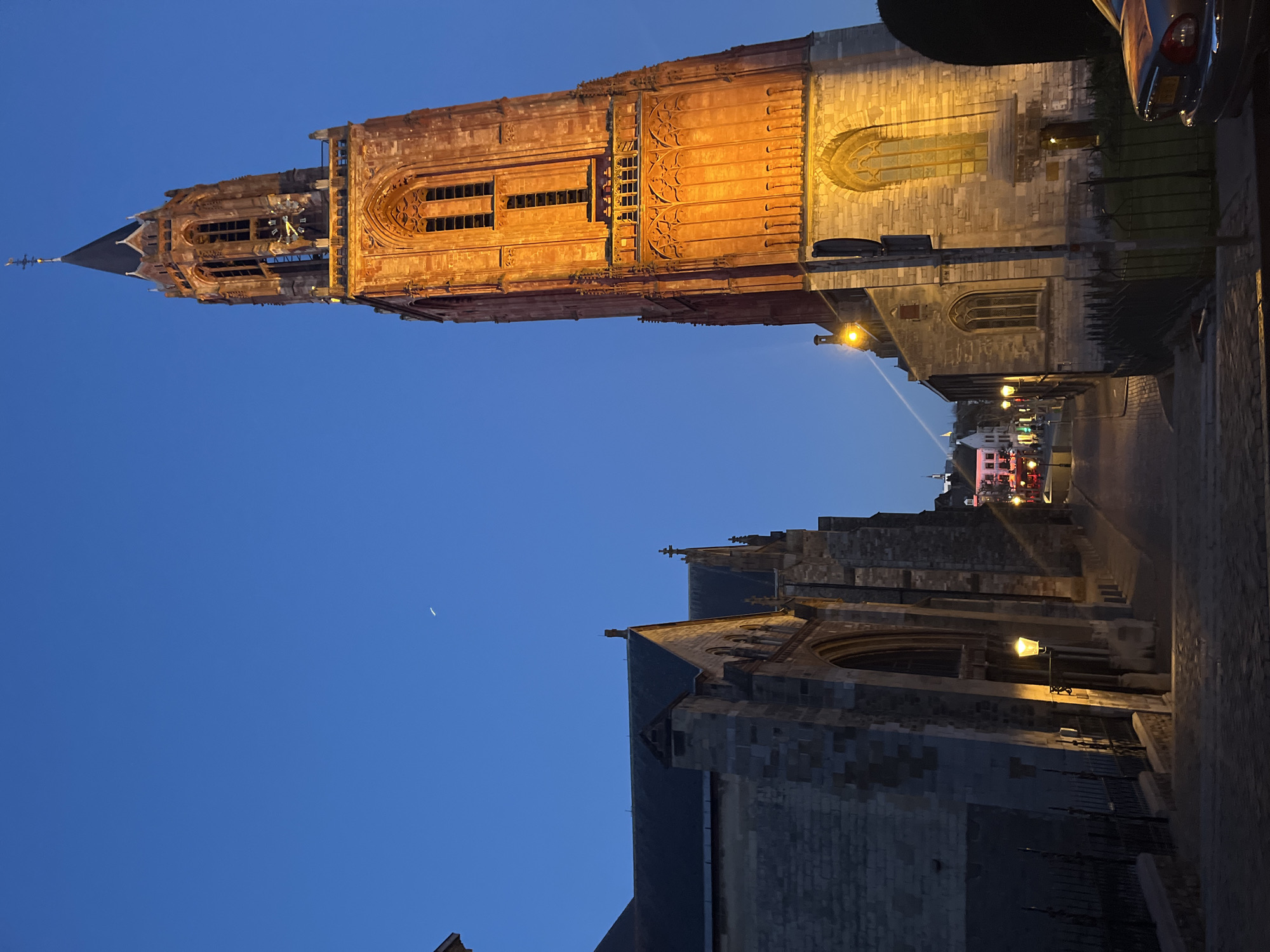 The image depicts a nighttime view of Maastricht, Netherlands, specifically featuring the Sint Janskerk (St. John's Church) and the adjacent Sint Servaasbasiliek (Basilica of Saint Servatius). The Sint Janskerk is a tall, slender, brick church tower illuminated by artificial light, contrasting with the dark blue sky. The Sint Servaasbasiliek, constructed of stone, is situated at the bottom of the frame, also partially lit. A crescent moon is visible in the upper left portion of the sky. A street runs between the two churches, with streetlights illuminating the path. Distant buildings with varied lighting are visible in the background, further down the street. There is a glimpse of a car's rear, and an indiscernible figure in the upper right corner of the image.