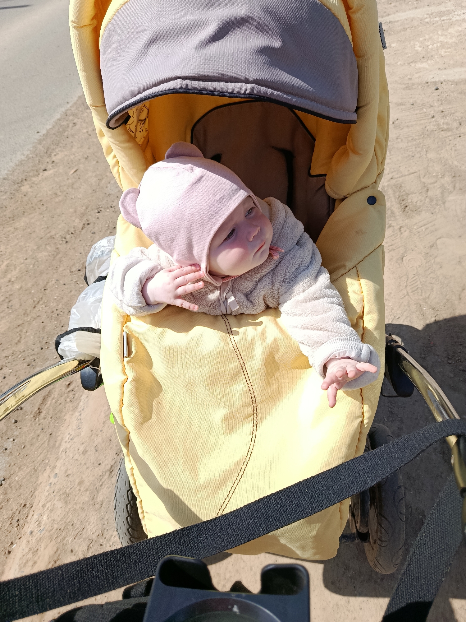 The photograph captures a baby seated in a yellow stroller, presumably outdoors in Фоки, Russia. The baby is wearing a pink cap with bear ears and a light-colored fleece jacket. The stroller's hood is partly open, revealing a grey interior. The stroller is positioned on a surface that appears to be a mix of dirt and pavement. A black strap is visible in the foreground.