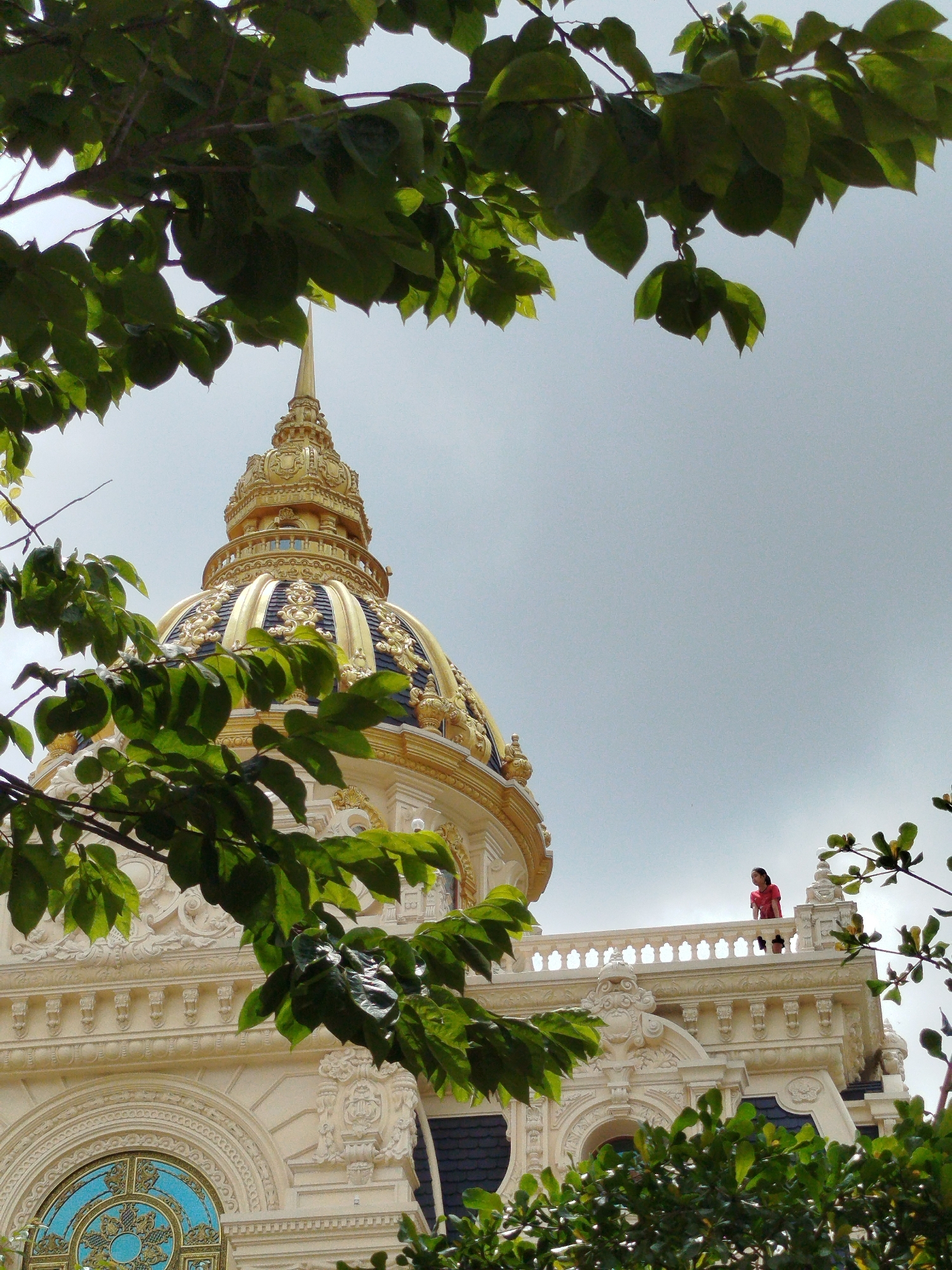 This image captures a view of the golden dome of the Thien Vuong pagoda, located in Da Lat, Vietnam. Lush green foliage from tree branches frames the shot, partially obscuring the ornate architecture. A person in a red shirt stands on a balcony of the pagoda, overlooking the surroundings. The sky above is overcast.