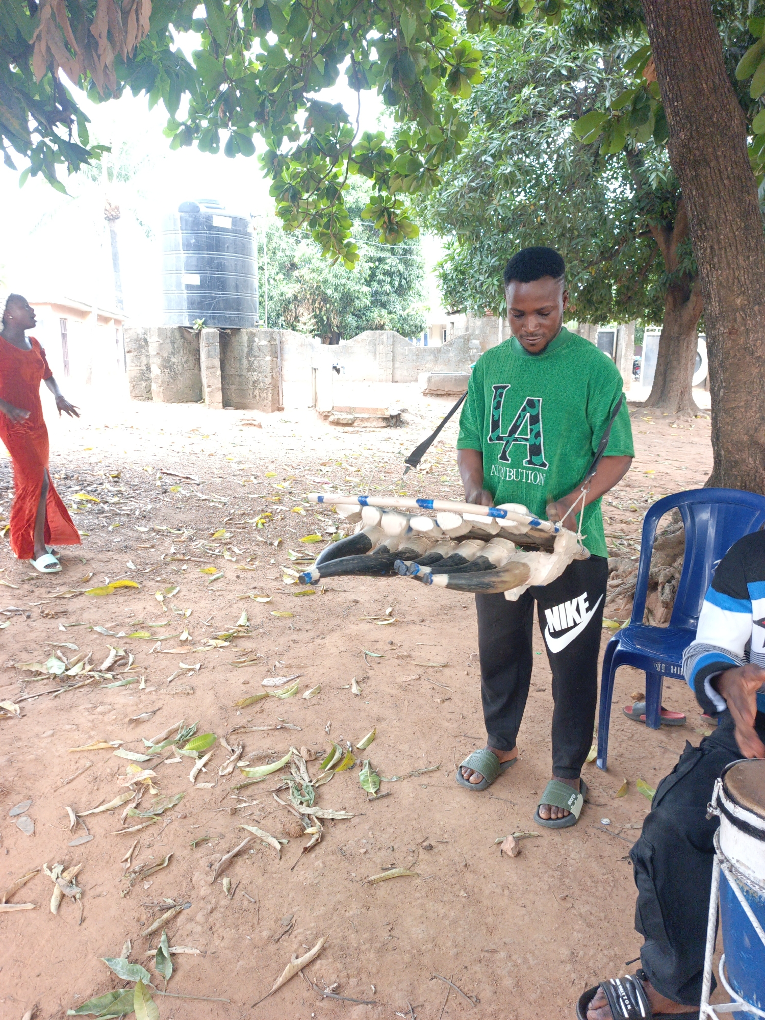 In Akwanga, Nigeria, the scene unfolds outdoors on a ground covered with fallen leaves. A man wearing a green "LA" shirt, black Nike pants, and green sandals is holding an object resembling a wooden carving of a horned animal. Beside him, a person sits near a blue plastic chair and a drum. In the background, a woman in a red dress stands near a water tank. Trees provide a canopy above.