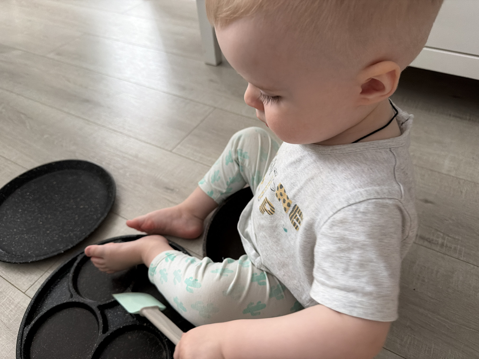A baby sits on a light gray, wood-patterned floor, likely indoors. The baby wears a light gray shirt with a giraffe design and light-colored pants with a green pattern. The baby's feet are near a black, round pan with several circular indentations and a detached black lid. The baby holds a light green spatula. The background includes a white object, possibly furniture. The location cannot be determined as Votkinsk, Russia, based on the provided visual information.