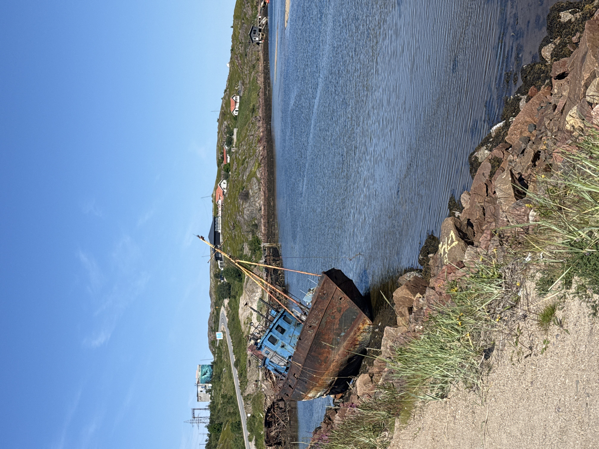 An abandoned, rusted fishing vessel is grounded on rocks in a bay in Teriberka, Russia. The vessel, predominantly brown with a blue cabin, is tilted significantly to its port side. The water in the bay is calm and blue, reflecting the clear, bright sky. Along the shoreline, red rocks and green grass are visible. In the background, a winding road leads to a cluster of houses situated on a grassy, rocky hillside. Further in the distance, industrial structures and power lines are present. The overall scene depicts a coastal landscape with elements of both nature and human habitation, marked by the presence of derelict maritime equipment.