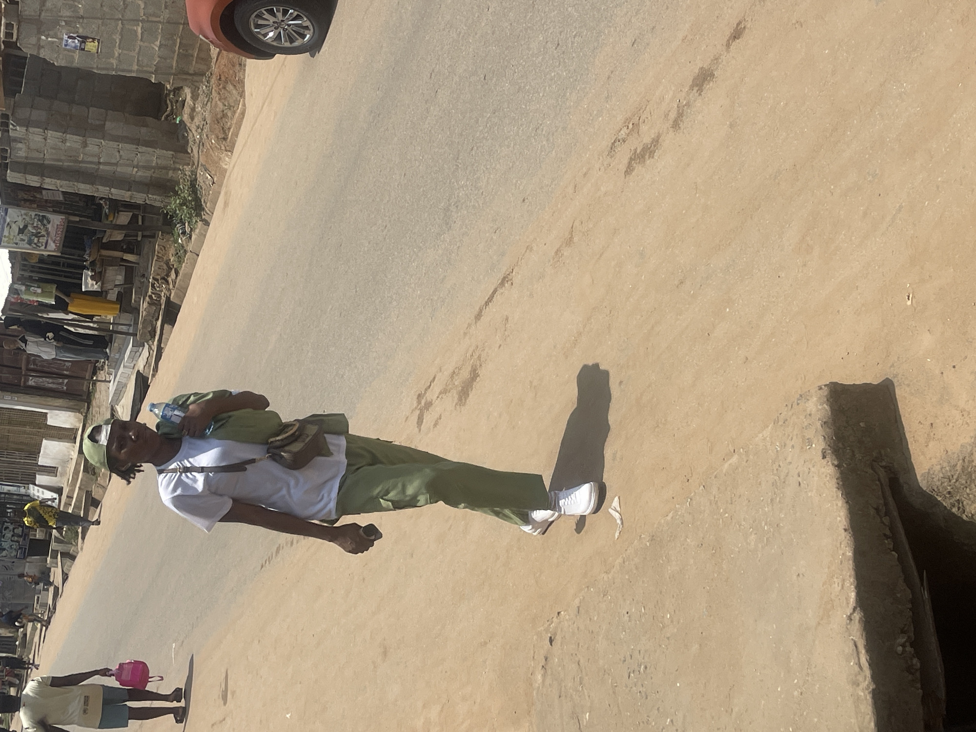 A young woman in a white t-shirt and green pants walks on a dusty road in Abuja, Nigeria. She wears a green cap and carries a water bottle and a bag over her shoulder. In the background, other people are walking and a car is parked on the side of the road. The scene appears to be in a busy urban area with buildings and shops lining the street.