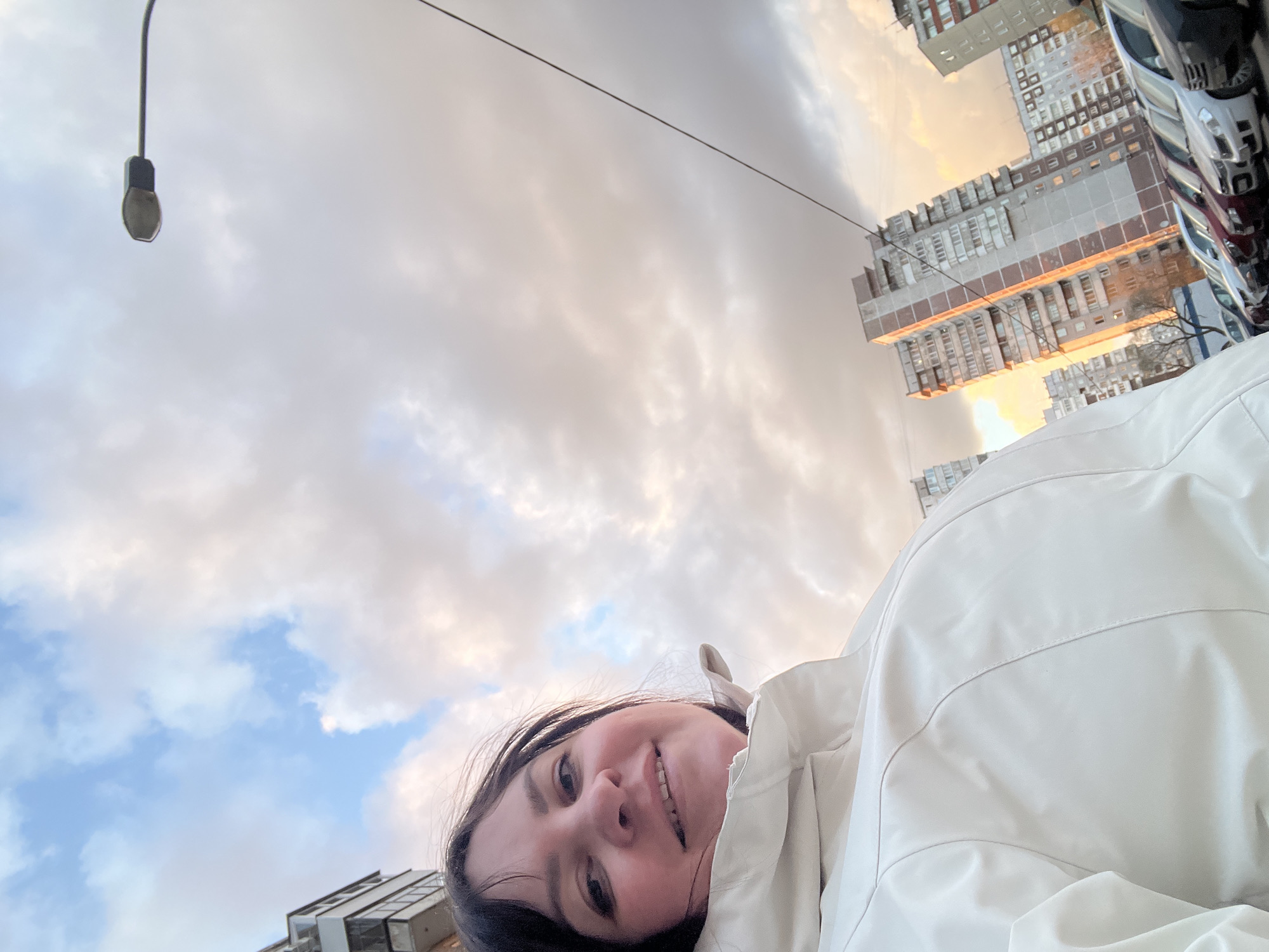 The image captures a low-angle outdoor selfie featuring a young woman smiling against a backdrop of an urban sky and buildings. The woman, with dark hair, is positioned in the lower right of the frame, looking directly at the camera with a cheerful, contented expression. She is wearing a light-colored, possibly white or light grey, padded jacket.
The sky above her is largely cloudy, displaying a blend of soft blue, white, and muted orange/pink tones, indicative of either sunrise or sunset. A street light fixture hangs from an overhead wire on the left side of the frame, with the wire extending across the sky.
In the background on the right, several multi-story residential buildings or apartment blocks are visible, characterized by numerous windows and balconies. Parts of parked vehicles can also be seen next to these structures. The architectural style and overall urban setting are consistent with the location context of Perm, Russia. There is no visible text in the image.