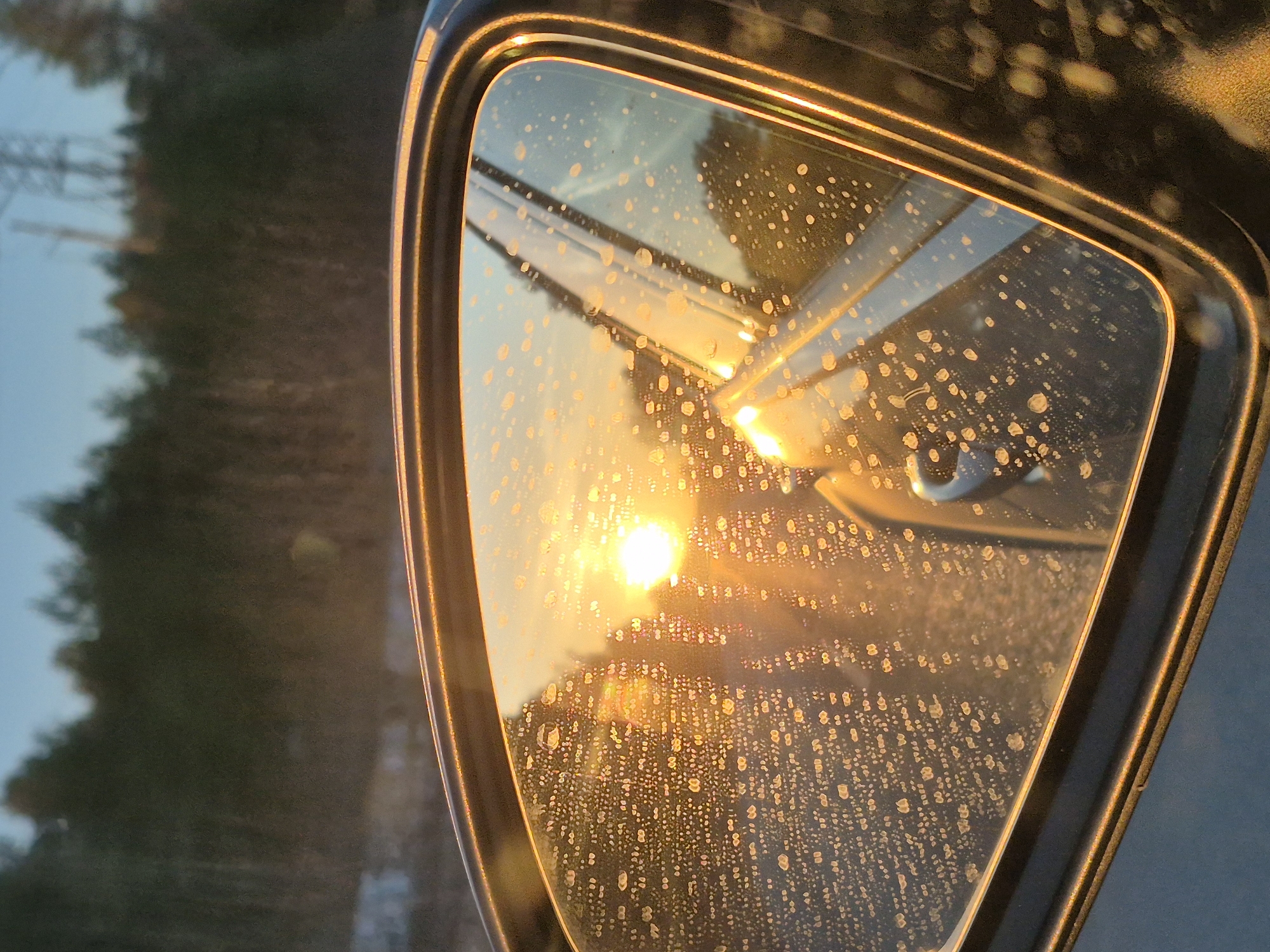 The image shows a car side mirror reflecting a sunset or sunrise. The mirror is covered in small water droplets. Reflected in the mirror is a bright orange sun or a reflection of the sun on a building. Behind the sun reflection, through the mirror, a blurred view of trees and utility poles or power lines is visible. The color palette is dominated by orange, yellow, and blue hues. No people are visible in the image. The location is unknown, the city of Noviy is not visible in the image.