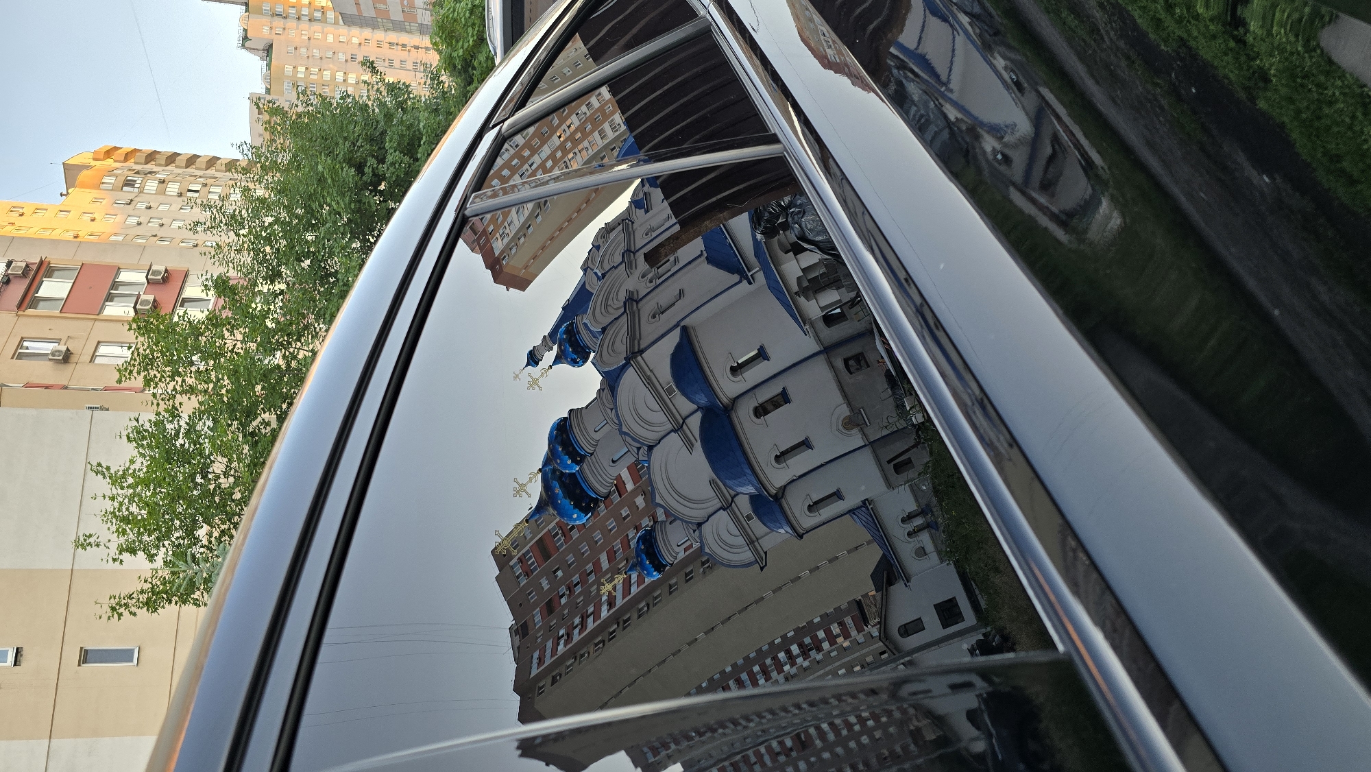 The image depicts a reflection of an Orthodox church with blue domes and white walls in the dark, polished surface of a car window. The church's reflection is prominent, showing multiple domes topped with golden crosses. Behind the church, tall apartment buildings with red and beige facades are visible, characteristic of urban architecture. Green trees are interspersed between the buildings and also appear in the reflection. The sky is a pale blue, suggesting it's either early morning or late afternoon. The car's window frame is also visible, made of chrome. The scene is set in an urban environment in Kyiv, Ukraine. There are no people clearly visible in the reflection or the scene itself.