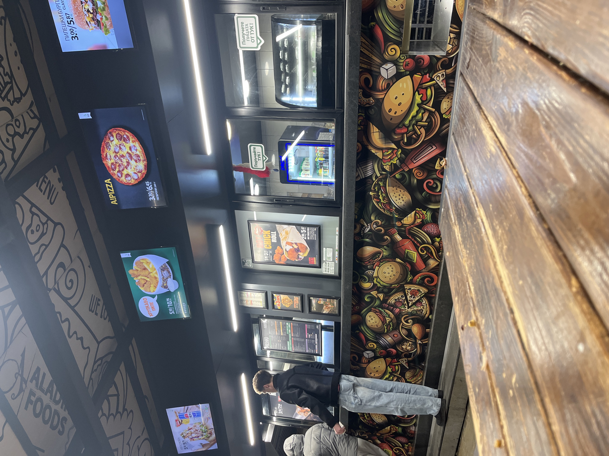 Here is a forensic description of the scene:
**Setting:** The image depicts the interior of a fast-food restaurant called "Aladdin Foods" with self-service kiosks. The decor includes a black and white illustrated wall with food themes and a brightly colored mural of various fast food items like burgers, pizzas, and hot dogs. There is a wooden structure adjacent to the wall.
**Subjects:** Two individuals are visible in the frame. One man is wearing a black jacket and light-colored jeans. He appears to be looking at the menu on the kiosks. The second person is mostly out of frame, wearing a light gray jacket and facing the same direction as the first man.
**Objects:**
* Multiple self-service kiosks displaying food items like pizza, chicken, and burgers with their prices.
* A food vending machine with food visible through the glass
* Signage in Bulgarian indicating order and pick-up locations ("Поръчайте ТУК!" and "Получете ПИЦАТА ОТ ТУК!").
* Screens with food advertisements and prices.
**Actions:**
* The primary action seems to be customers interacting with the self-service kiosks, likely placing orders.
**Location:** The location is the interior of Aladdin Foods in Unknown, Unknown. Specific city and country are unspecified.
**Environment:** The environment appears to be a clean, modern fast-food establishment with a focus on self-service ordering and pick-up.