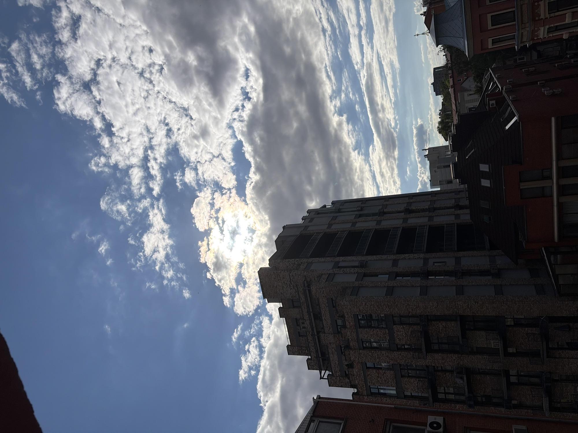 The image shows a view of urban buildings under a partly cloudy sky, with the sun partially obscured by clouds, creating a halo effect. The architecture suggests a European city, possibly Dnipro, Ukraine, given the provided context. The foreground features the upper floors of several buildings, including a prominent brick high-rise with dark-framed windows and balconies. To its left is a portion of another brick building with a visible air conditioning unit. In the background, a mix of residential and commercial structures can be seen, with pitched roofs and varied facades. The sky is a mix of bright blue and white, with scattered cumulus clouds. There are no visible people or vehicles. The overall impression is that of a city scene during daylight, with atmospheric conditions affecting the lighting.