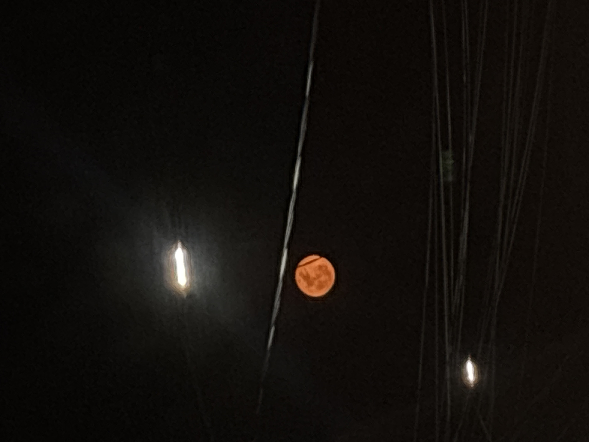 The image, captured at night, features a reddish-orange full moon partially obscured by what appear to be electrical wires or thin tree branches. To the left of the moon, a vertical streak of bright light, possibly from a street lamp, cuts through the darkness. There is a second streak of light in the lower right corner of the image, also likely from a streetlamp. The environment is dark, with little visible detail in the background. The image suggests an urban or suburban setting where utility lines are present, but without additional context, the location in Cuernavaca, Mexico is indeterminate.
