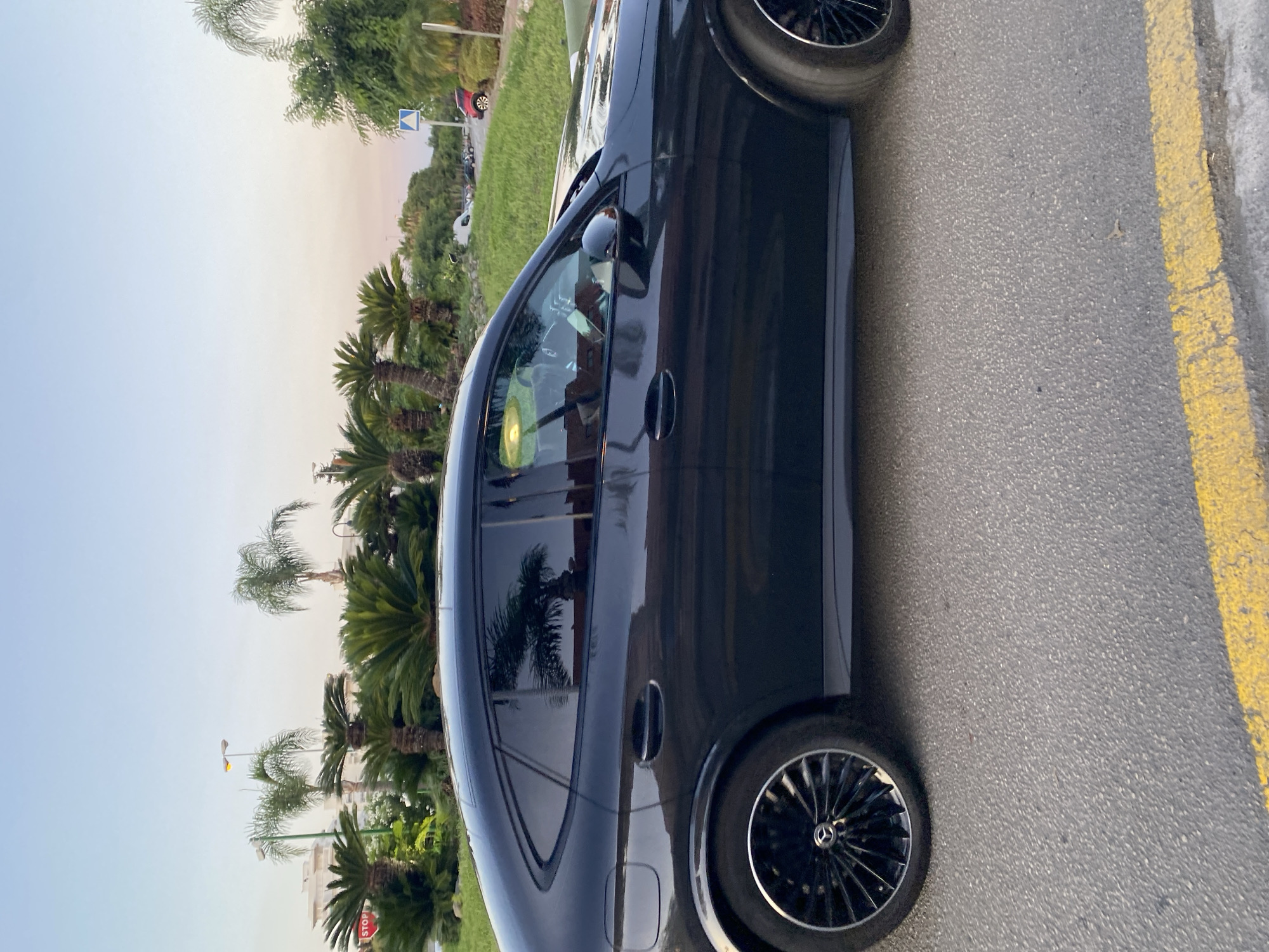The image shows a dark-colored Mercedes-Benz coupe parked on a street, likely in Resinera-voladilla, Spain, given the presence of palm trees and the overall aesthetic. The sky is a pale blue, transitioning to a soft pink and orange hue near the horizon, indicating it's either dawn or dusk. The car is the primary subject, its sleek profile and tinted windows are visible. Reflections of palm trees are discernible on the car's windows. The street is paved, with a yellow line marking the edge of the road. In the background, lush green vegetation, including various types of palm trees and bushes, lines the street. There are also buildings and streetlights visible in the distance, suggesting an urban or suburban setting. A red car is parked further down the street. The scene is quiet and serene, with no people clearly visible in or around the car.