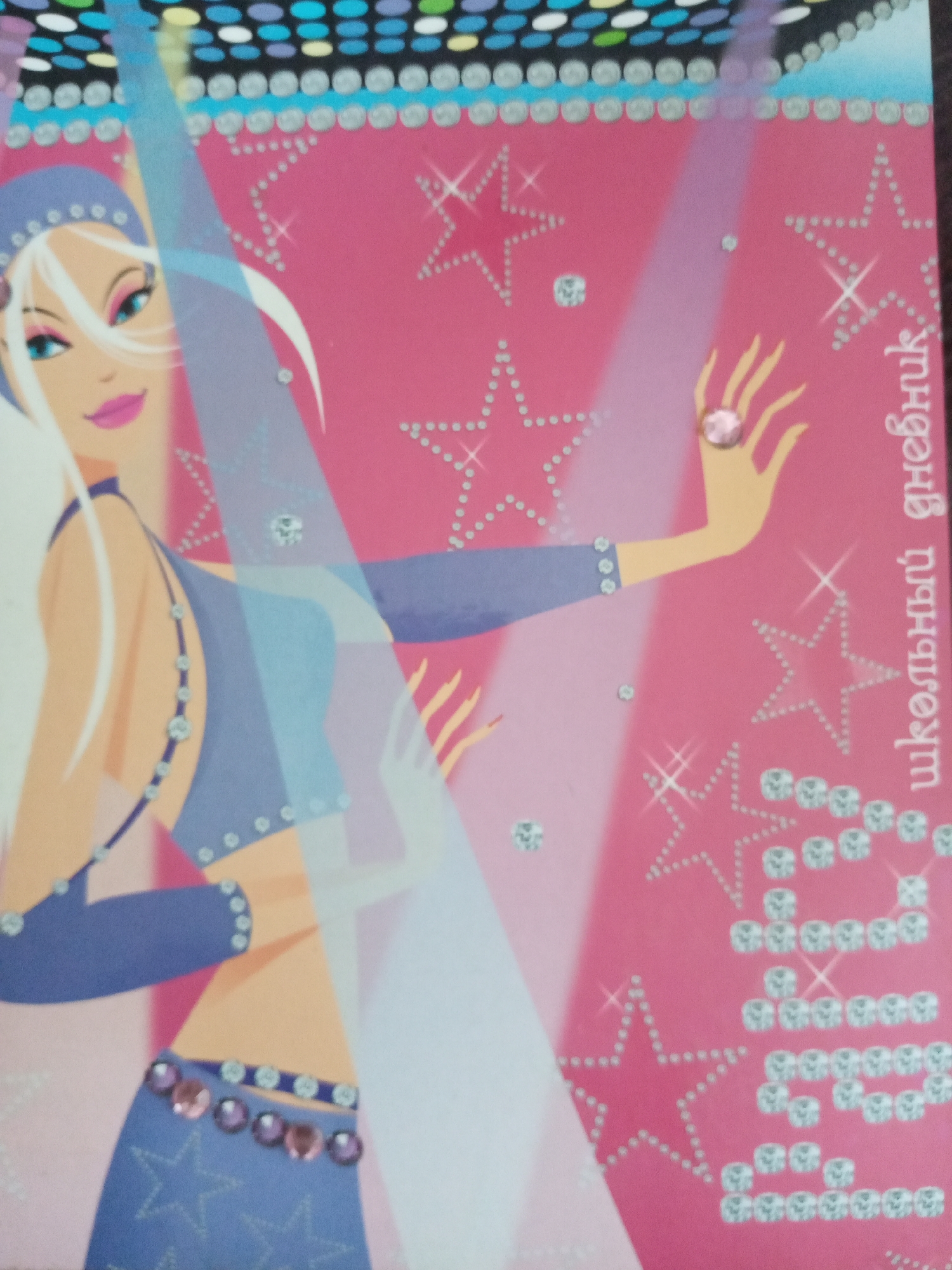 The image shows a close-up of a notebook cover with a cartoonish illustration of a fair-skinned, blonde-haired female figure in a blue outfit with various jewel-like embellishments. The background is pink with dotted stars. Russian text is visible on the right side, spelling "школьный дневник" (shkol'nyy dnevnik), meaning "school diary."
The image does not depict a scene or location in the city of Elektrostal, Russia.