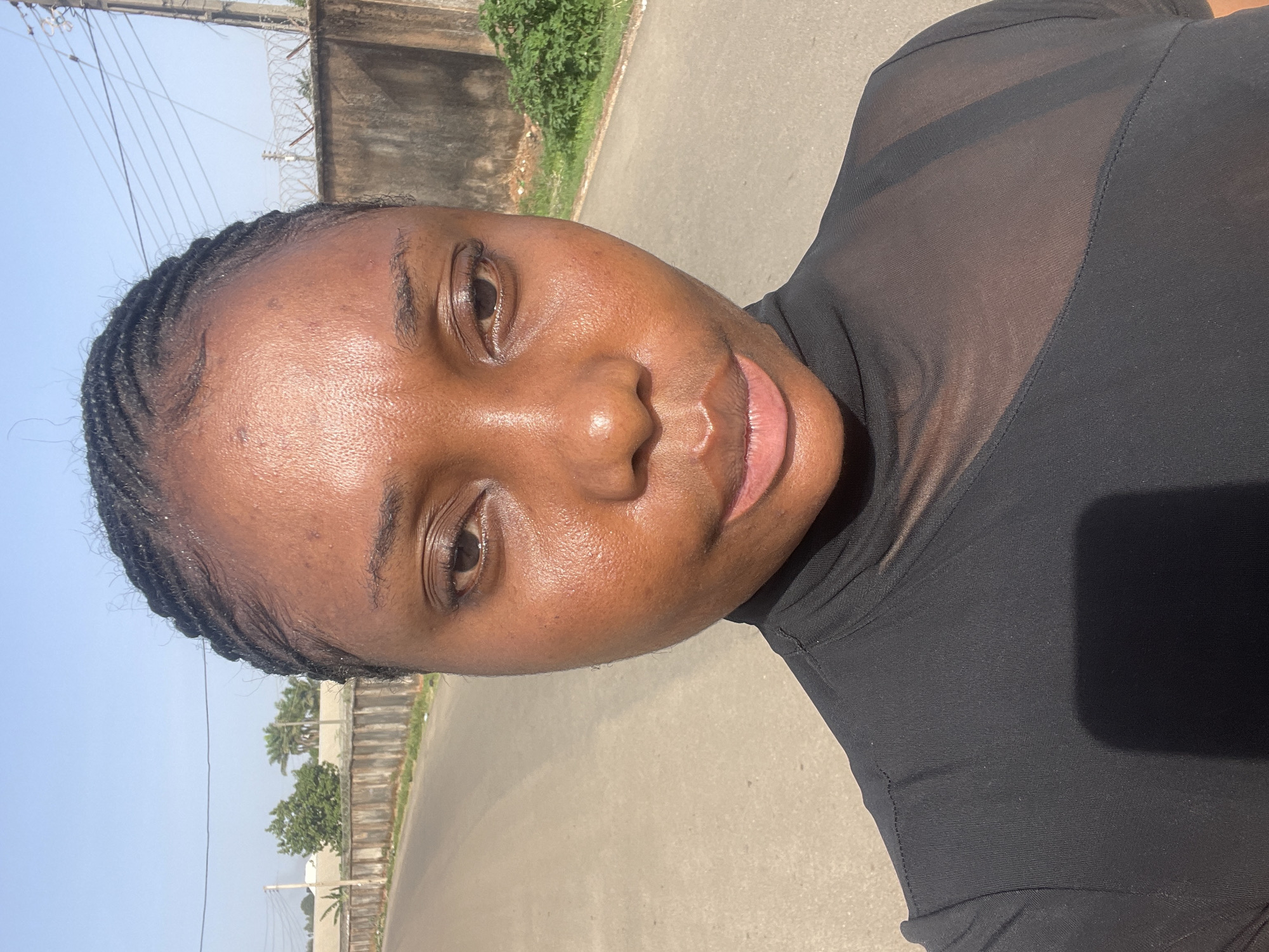 The image displays an outdoor, angled portrait of a young Black woman, likely taken as a selfie. She is looking directly at the camera with a calm, neutral expression, possibly a subtle, gentle smile. Her hair is styled in neat braids pulled back from her face. Her skin, well-lit by bright sunlight, shows a natural sheen and texture.
She is wearing a black top featuring sheer or mesh panels on the upper chest and shoulders, through which a black bra strap is visible. The bright, direct sunlight creates noticeable highlights on her skin and suggests the photograph was taken during midday or early afternoon.
The background depicts a sunny outdoor environment consistent with Jos, Nigeria. A paved road or path stretches behind her, flanked by a concrete wall or fence. Beyond this wall, there are sparse trees, including palm trees, and bushes, along with utility poles and wires, all set against a clear, light blue sky. There is no visible text in the image.