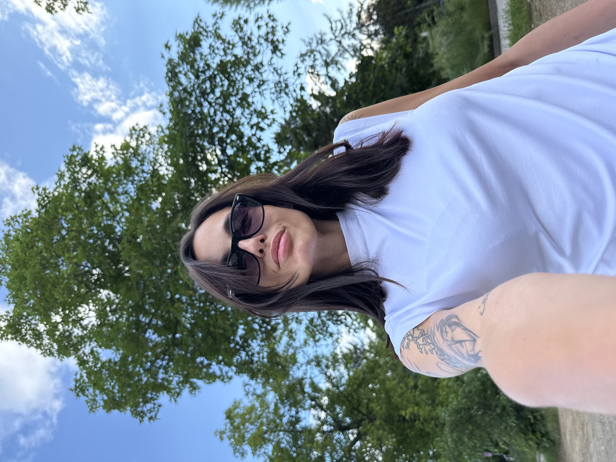 A woman wearing sunglasses and a white t-shirt is photographed from a low angle, with trees and a blue sky visible in the background. The woman has dark hair and a tattoo on her arm. The photograph appears to be taken outdoors. The specific location in the city of Pleven, Bulgaria is not discernible from the image.