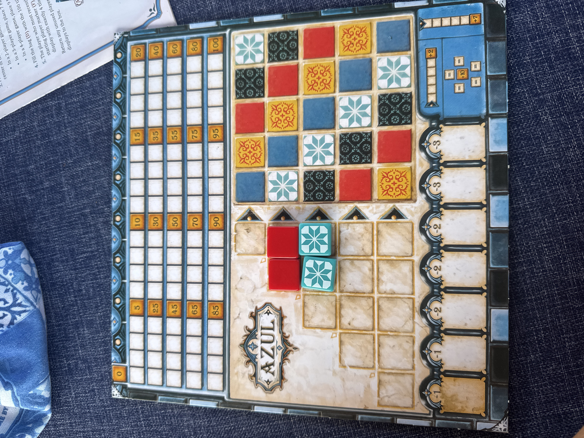 The image displays a tabletop game board, identified as "AZUL", set on a textured blue fabric surface, possibly denim or a similar material. The game board features a grid of squares for tile placement and a scoring track. Several tiles are visible on the board: two red tiles and two turquoise tiles with a star pattern are in the lower-left section of the player's board. A portion of a rulebook with text and diagrams is visible in the upper right corner of the image. The game board itself has a decorative border and an area for storing tiles. The lighting is even, and the focus is on the game board and its components. There are no people or animals present in the image. The location of Kuta Selatan, Indonesia is not determinable from the visual information provided in the image.