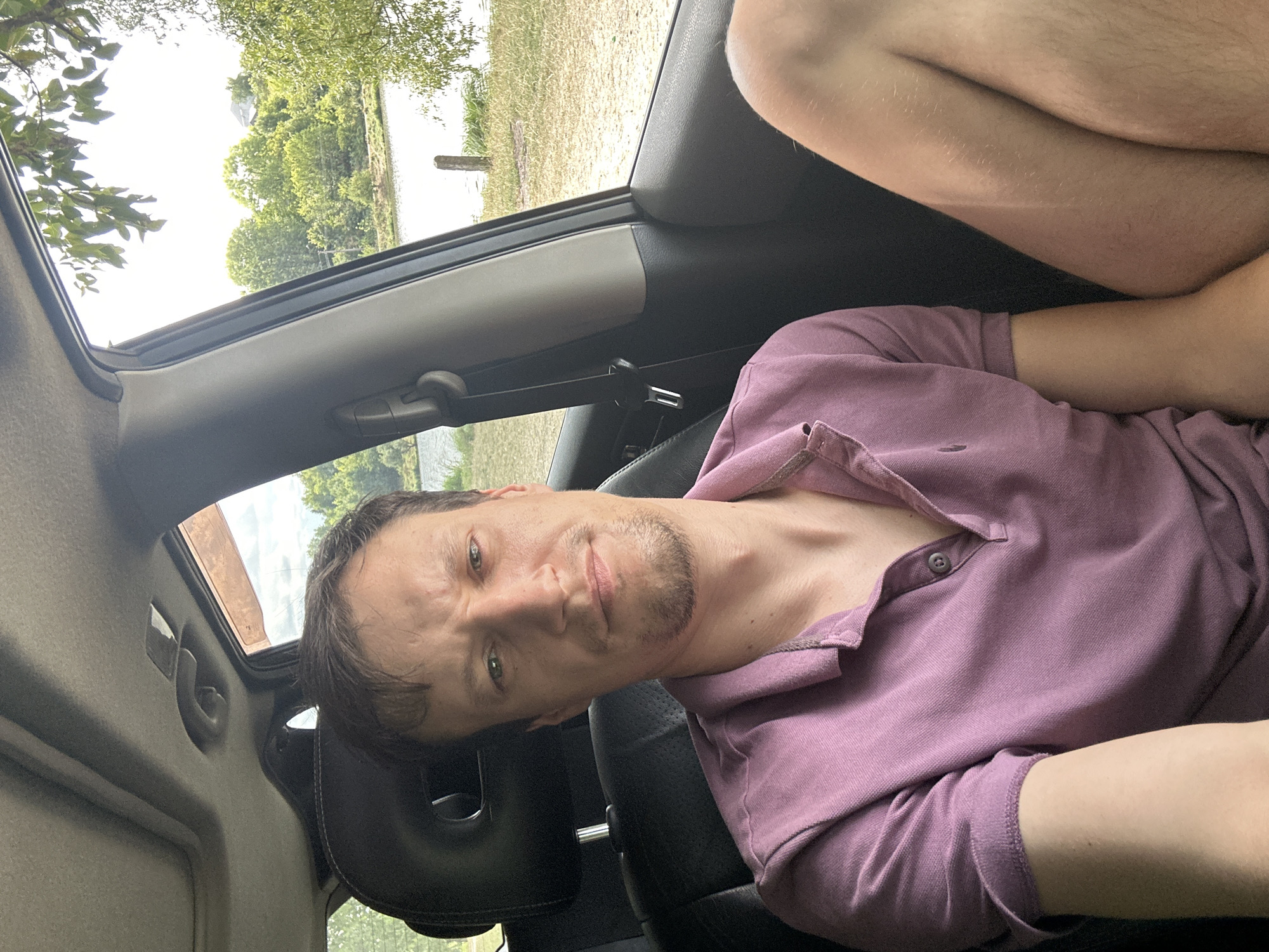 A man with short brown hair and a short beard is sitting in the driver's seat of a car, looking at the camera. He is wearing a purple polo shirt. Through the open car window, a body of water, trees, and a bright sky are visible. The man's legs are also visible in the foreground. The scene appears to be a scenic location, possibly a park or roadside near a body of water in or around Makiv, Ukraine, given the context.