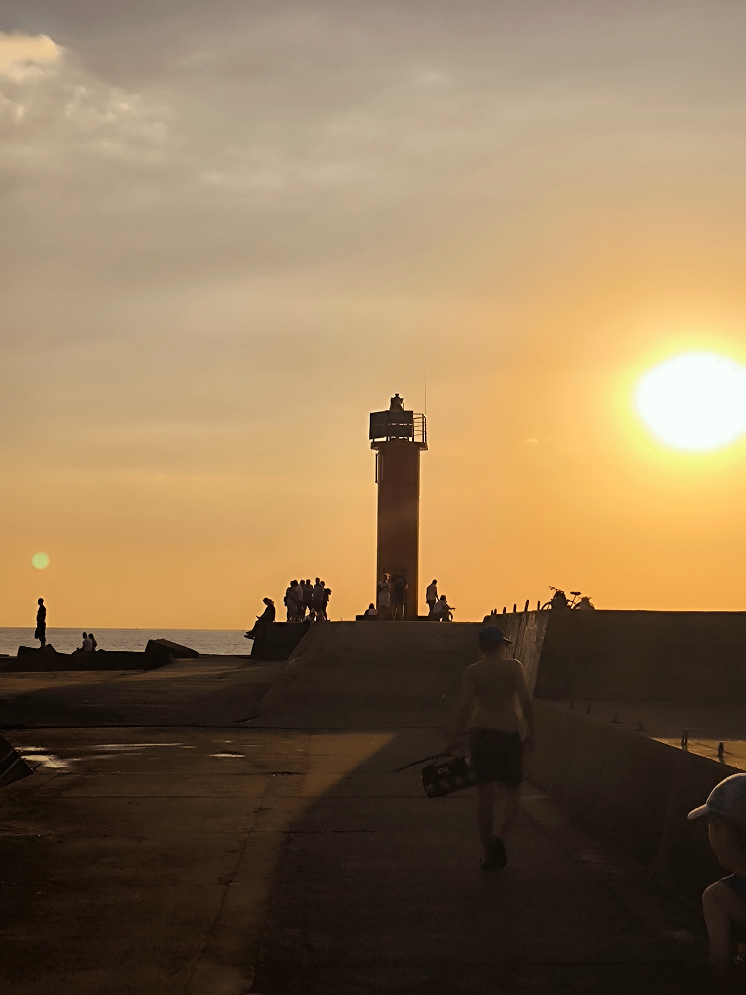 The image depicts a sunset over the Baltic Sea at Daugava, Riga, Latvia. A lighthouse stands prominently in the middle ground, silhouetted against the bright orange sky. Several people are visible on the pier, some standing and observing the sunset, while others are seated. In the foreground, a person walks away from the camera, carrying a bag. The sun is a bright disc low on the horizon, casting long shadows across the concrete pier. The water is calm, reflecting the warm hues of the sunset.
