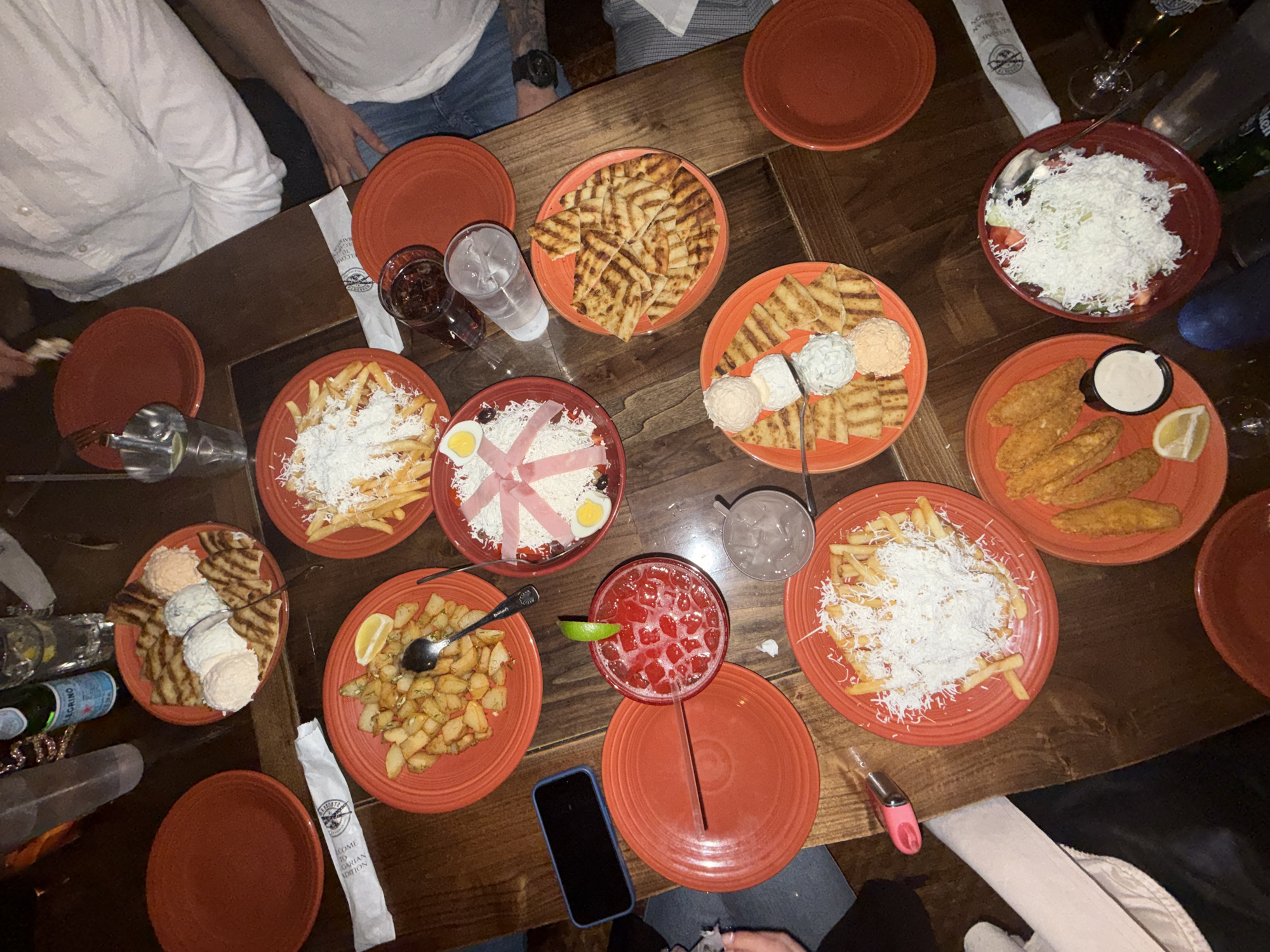 A high-angle shot depicts a wooden table laden with orange-red plates of food and beverages. The setting appears to be a restaurant, with portions of people visible around the edges of the frame. There are no distinguishing characteristics to allow the determination of the location in Des Plaines, United States of America.
Various dishes are arrayed across the table: one plate contains grilled pita bread; another contains fried potatoes topped with grated white cheese; a third has fried potatoes with a lemon wedge; another plate has salad with shredded white cheese; a plate has fried fish strips with a dipping sauce and a lemon wedge; another has a dish containing a pink cocktail. There are multiple empty plates also visible. Additional objects include a glass of water, a glass of dark liquid, a smartphone, a bottle of sparkling water, silverware, and a pink cigarette lighter. Visible partial people include those wearing light-colored shirts and jeans, with a tattoo visible on an arm.