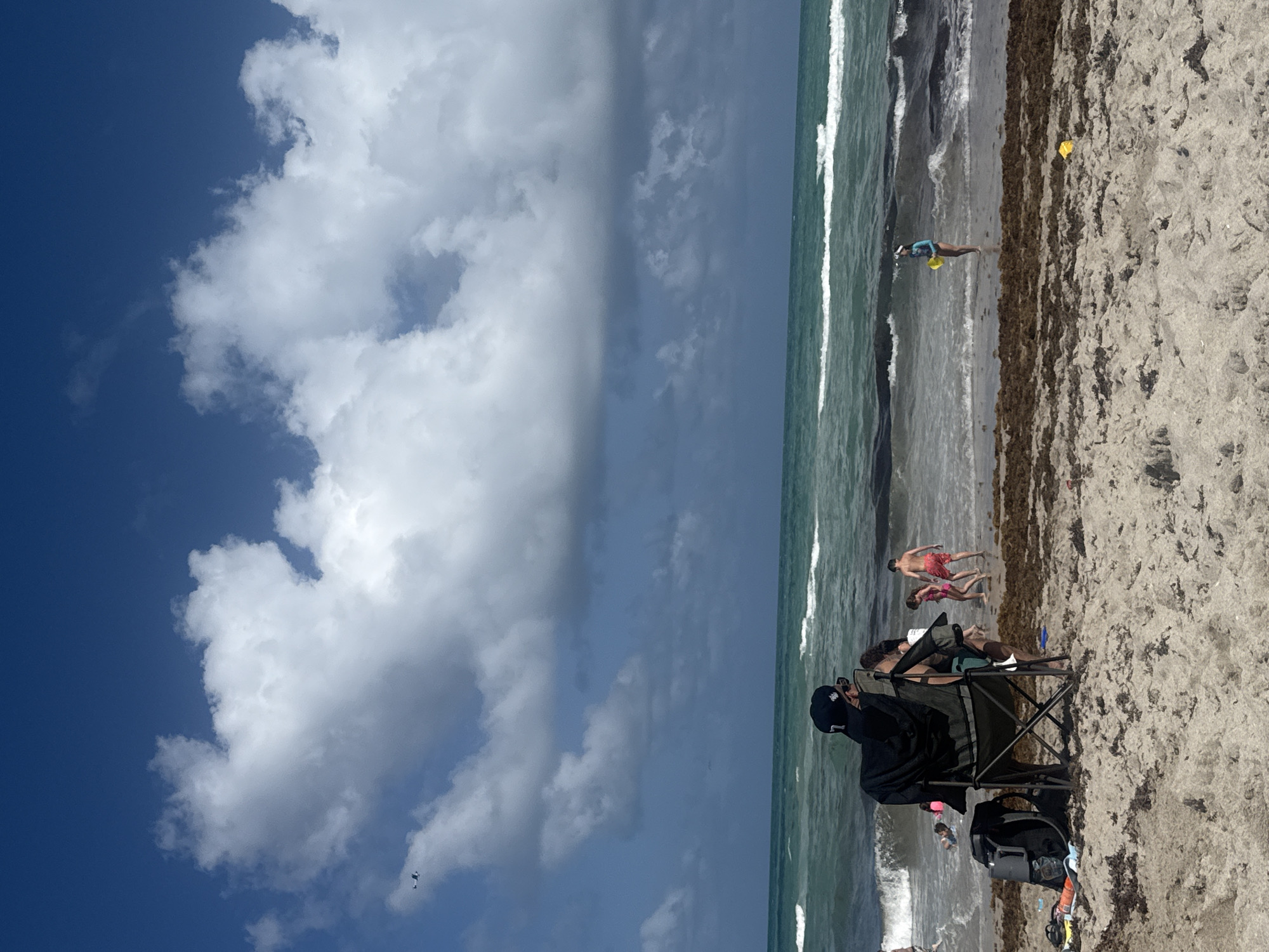 The image depicts a beach scene in Hollywood, Florida, United States. The sky is partly cloudy with a mix of blue and grey tones. The ocean meets the shore, with waves visible near the coastline and seaweed scattered on the sand. Several people are present on the beach, some are wading in the water, others are walking along the shoreline, and one person is sitting in a folding chair. Objects such as a backpack and other beach equipment are near the seated person. The overall setting is a typical beach day, with people engaged in various recreational activities.