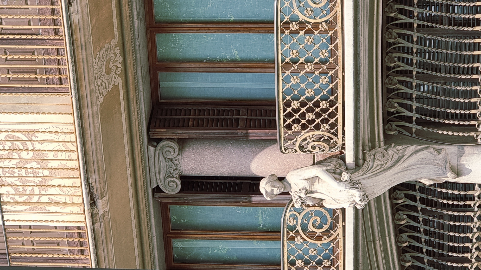 The image captures a portion of the facade of a building in Olot, Spain, specifically the Casa Balsach, located on Carrer del Bisbe Lorenzana. The focus is on architectural details and a sculpted figure.
In the foreground, a white stone sculpture of a woman is prominently displayed, appearing to emerge from the building's structure. She is depicted in a flowing gown, holding a bouquet of flowers. Her posture suggests a graceful, almost ethereal presence. The sculpture is attached to the building at what appears to be the lower level of a balcony.
Above the sculpture, a decorative balcony railing made of ornate metalwork with floral motifs is visible. This railing spans across a set of tall windows with wooden frames and louvered shutters, some of which are partially open. The windows are covered by light blue curtains.
Behind the balcony and windows, a large, fluted column with a classical Ionic capital rises vertically, partially obscuring the view of the windows. The column is made of a speckled, pinkish-grey stone. The upper part of the building features a cornice with decorative carvings and a second balcony with a similar railing to the one below. The overall impression is one of detailed craftsmanship and artistic embellishment on the building's facade. The lighting suggests a daytime scene, with sunlight casting shadows and highlighting the textures of the materials.