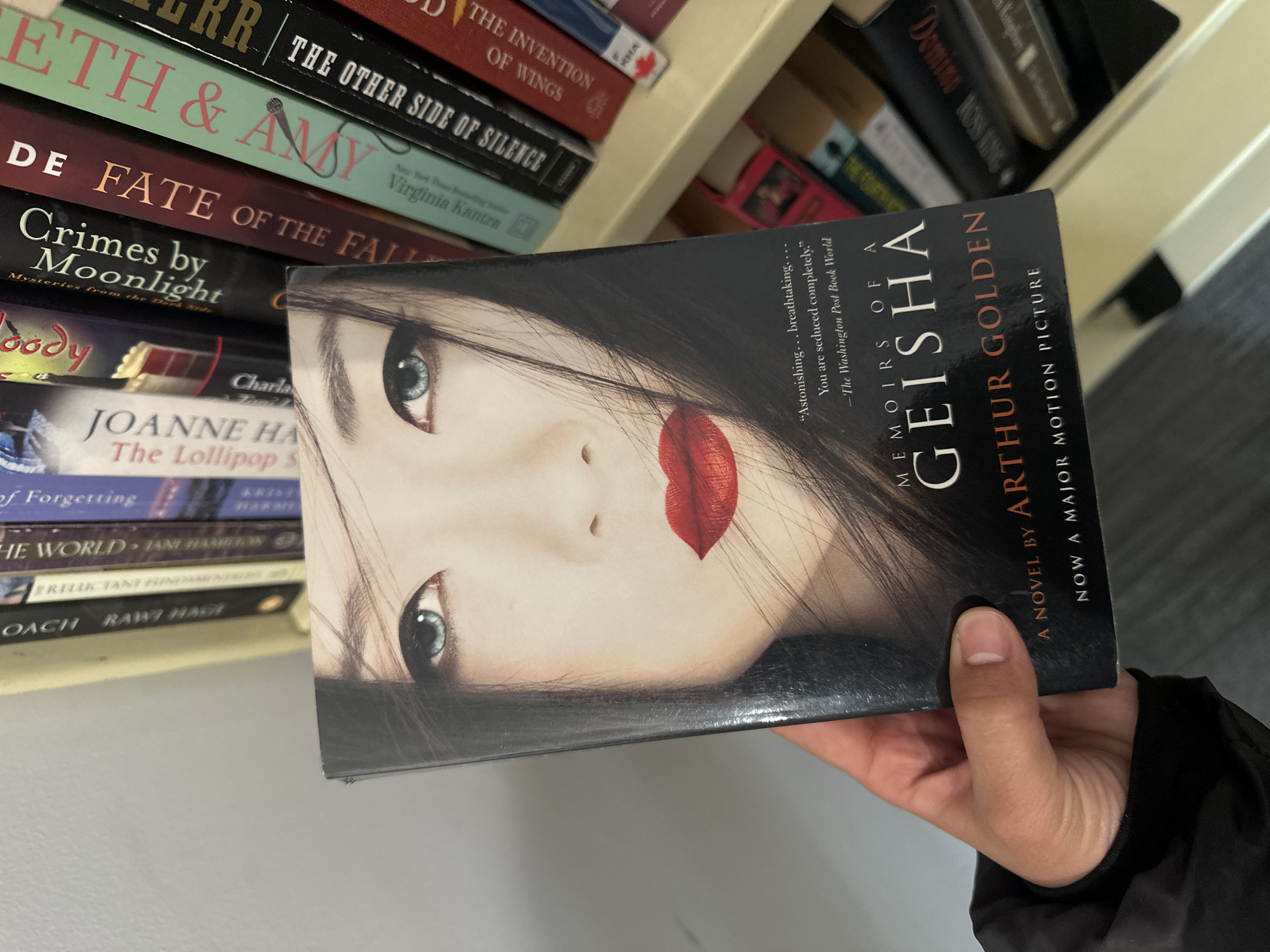 A person's right hand holds the novel "Memoirs of a Geisha" by Arthur Golden, published as "Now a Major Motion Picture," in an indoor setting, possibly a bookstore or library, located in New Westminster, Canada. The book's cover features a woman's face with dark hair, light blue eyes, and prominent red lips. A quote on the cover reads, "'Astonishing... breathtaking. You are seduced completely.' -The Washington Post Book World." The hand, with light skin and short fingernails, wears a dark long-sleeved garment. In the background, slightly out of focus, are several bookshelves filled with other books. Visible titles on the shelves include "The Invention of Wings," "The Other Side of Silence," "Eth & Amy" by Virginia Kantra, "De Fate of the Fall," "Crimes by Moonlight," "Joanne Ha The Lollipop S," "of Forgetting," "The World," "Oach Raw Hagi," and "Domino Rossing." The lighting suggests an artificial indoor light source.