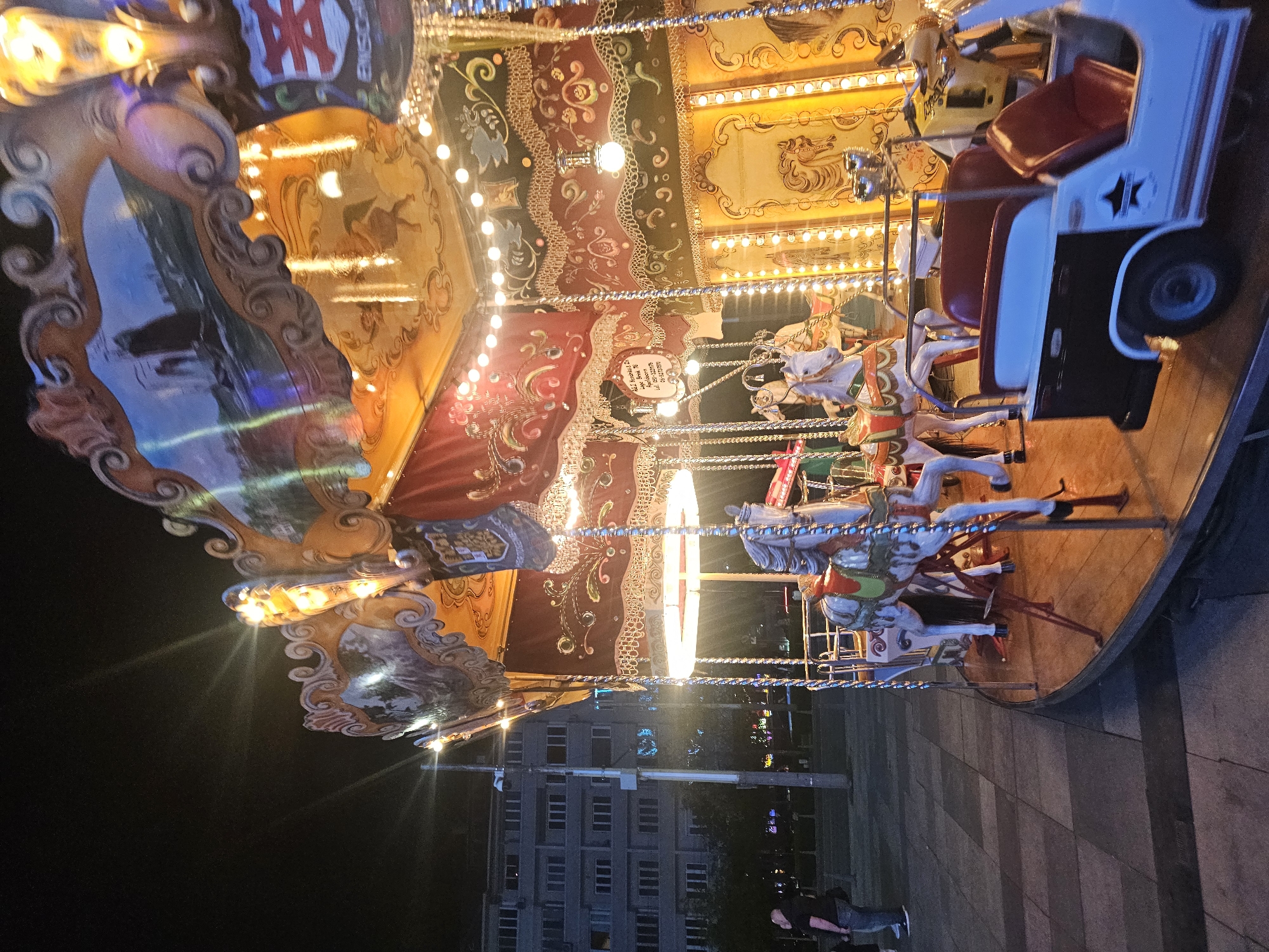 The image is of a carousel at night, with an illuminated canopy. The carousel has horses and a small vehicle for riders. There are decorative paintings on the carousel canopy, with a mix of red, gold, and other colors. There are buildings in the background, and two people are visible in the lower right corner. The location is Sofia, Bulgaria.