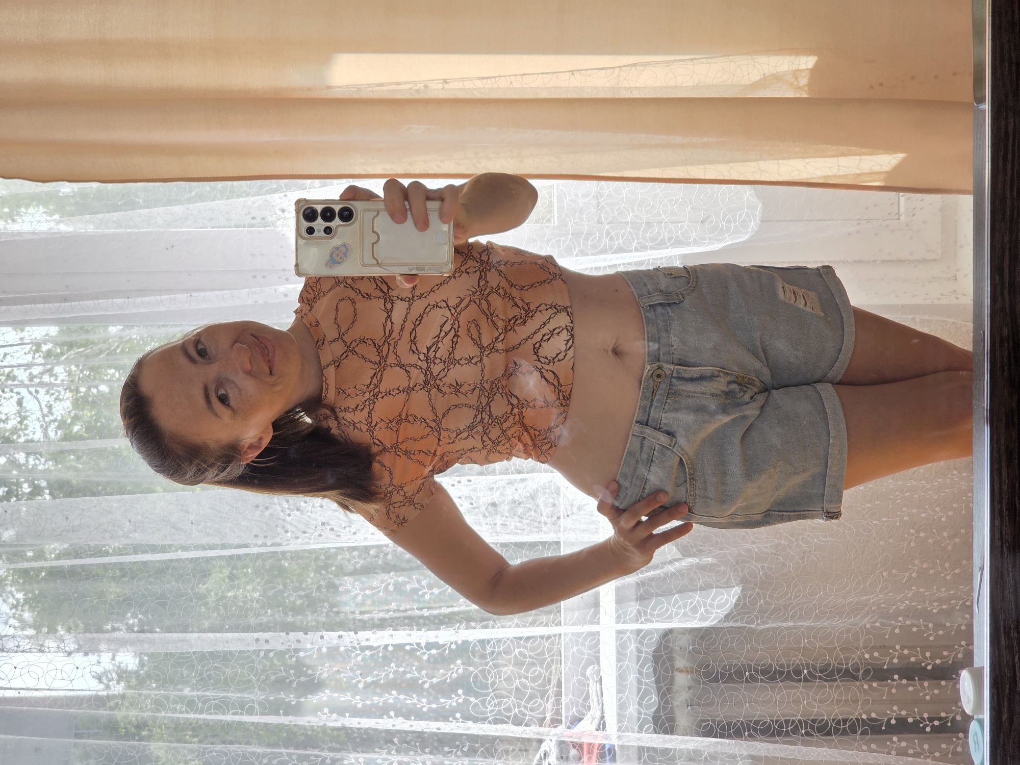 A full-body mirror selfie shows a woman standing in front of sheer white curtains, with a light peach curtain on the right. She has brown hair pulled back in a ponytail and is wearing an orange crop top with a dark chain-link pattern and light-wash denim shorts. Her right hand is on her hip, and she is holding a smartphone in her left hand, angled to capture the reflection. The background reveals trees and sunlight filtering through the curtains, suggesting a daytime setting. The environment appears to be an indoor space with a window. The woman is looking directly at the camera with a slight smile. No specific location within Chaikovsky, Russia, can be identified from the image.