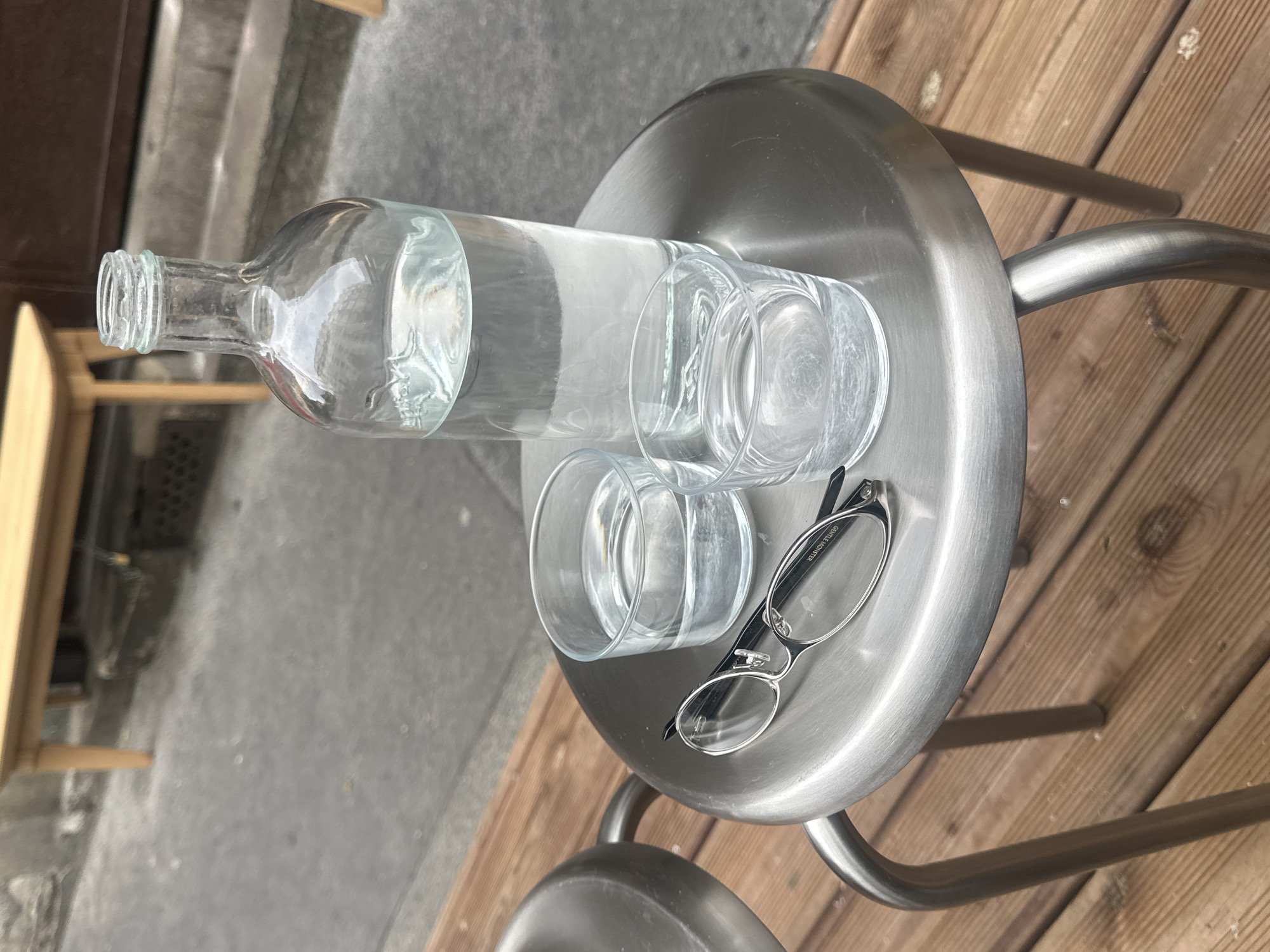 On a small, round, metallic outdoor table in Paris, France, there's a clear glass bottle partially filled with water, along with two empty clear glasses, and a pair of black-and-silver rimmed eyeglasses. The table, situated on a wooden deck, is positioned near a building exterior and sidewalk. The bottle is lying horizontally, with its neck facing left, while the glasses and eyeglasses are arranged to the right of it.