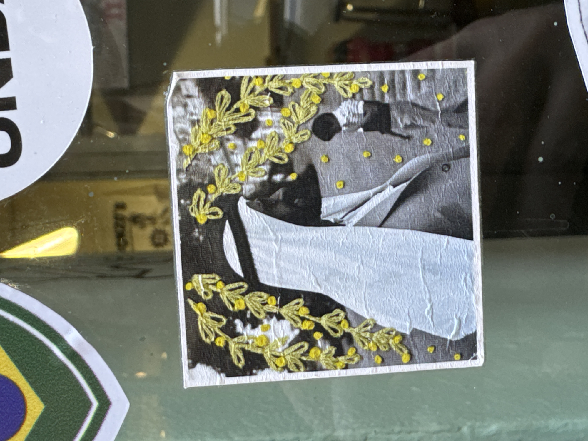 A close-up shows a square sticker adhered to a surface, possibly glass. The sticker features a black and white image of what appears to be a person or persons in formal attire, overlaid with yellow embroidered or drawn floral decorations. The paper of the sticker is creased and worn. The surface the sticker is adhered to has other stickers, including one partially visible with white lettering on a black background and another with the colors of the Brazilian flag. The environment appears to be indoors, with reflections visible in the glass surface. There is no information in the image to confirm it is taken in Milan, Italy.