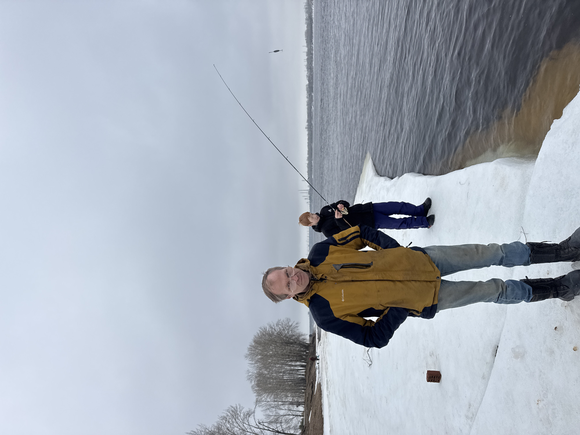 The image shows two people fishing on the shore of a body of water, possibly the Kama River. The location is likely in Chaikovsky, Russia. The foreground features a man in a brown and blue jacket, jeans, and black boots standing on a snow-covered surface. Behind him, a second person is fishing with a rod, lying on the snow. The sky is overcast, and the water is gray. Trees are visible near the shore. A fishing lure is seen suspended in the air. The bank appears to be icy.