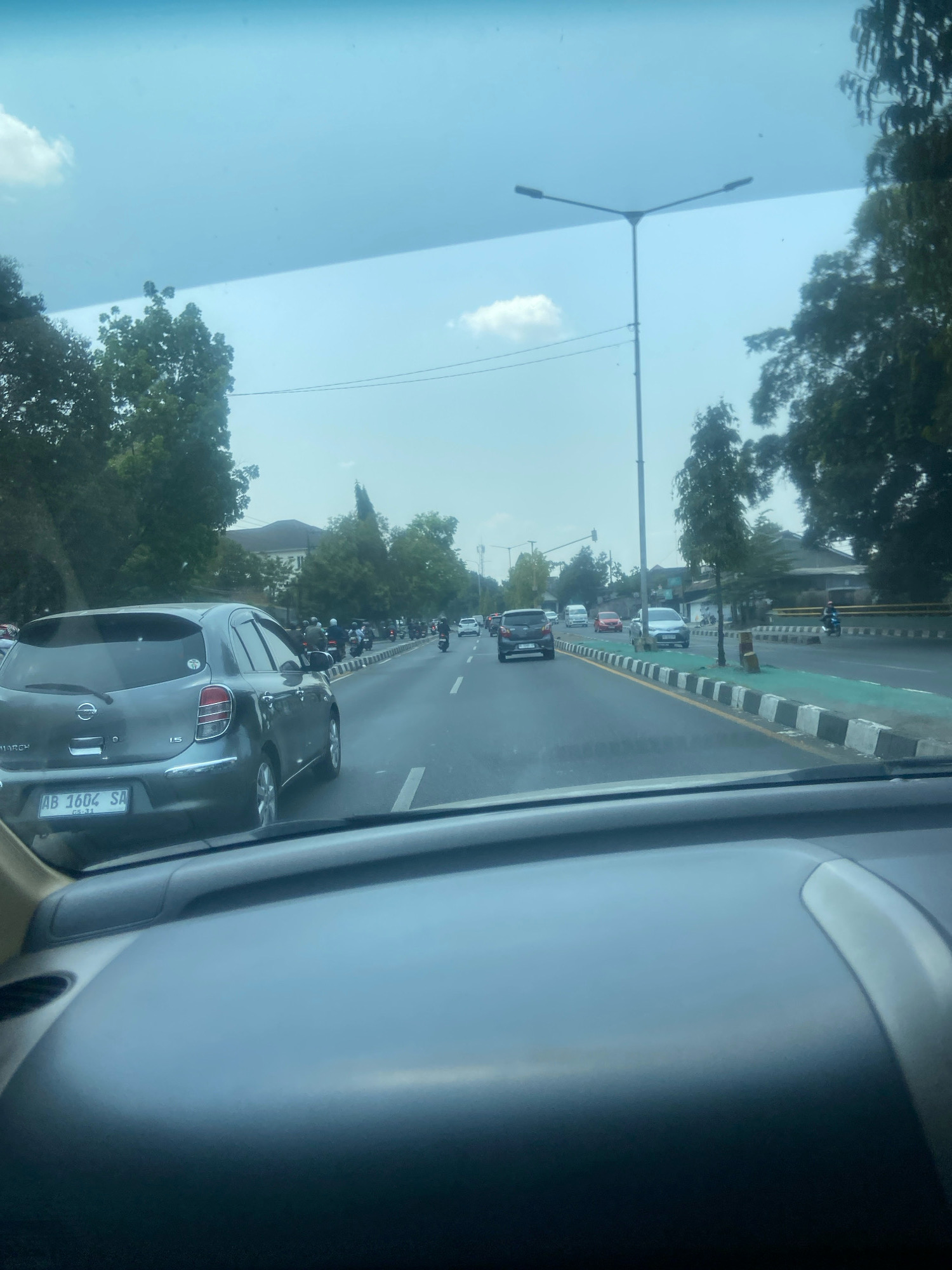 The image depicts a street scene in Ngaglik, Indonesia, viewed from inside a vehicle. The foreground shows the dashboard of the vehicle. In the middle ground, a grey Nissan March car with license plate "AB 1602 SA" is parked. Beyond that, a busy road is visible with numerous motorcycles and cars in traffic. Trees line the sides of the road, and a street light stands tall. The sky is overcast with some clouds. The overall scene suggests a typical day in an urban or semi-urban environment with moderate traffic.
