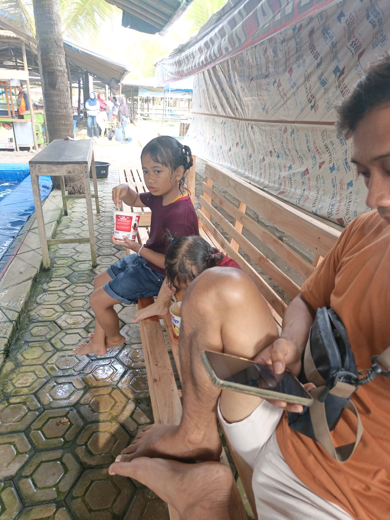 In Tambakmulyo, Indonesia, two young girls and an adult are seated on a wooden bench near a pool. The older girl, wearing a maroon shirt and denim shorts, holds a white container with a red lid, possibly food. The younger child is partially visible next to her. The adult, wearing an orange shirt, holds a smartphone and a dark bag is slung over their shoulder. The ground is paved with hexagonal tiles, wet and reflecting light. In the background, palm trees and a covered structure suggest an outdoor recreational area or market. A blue pool is visible to the left of the bench.
