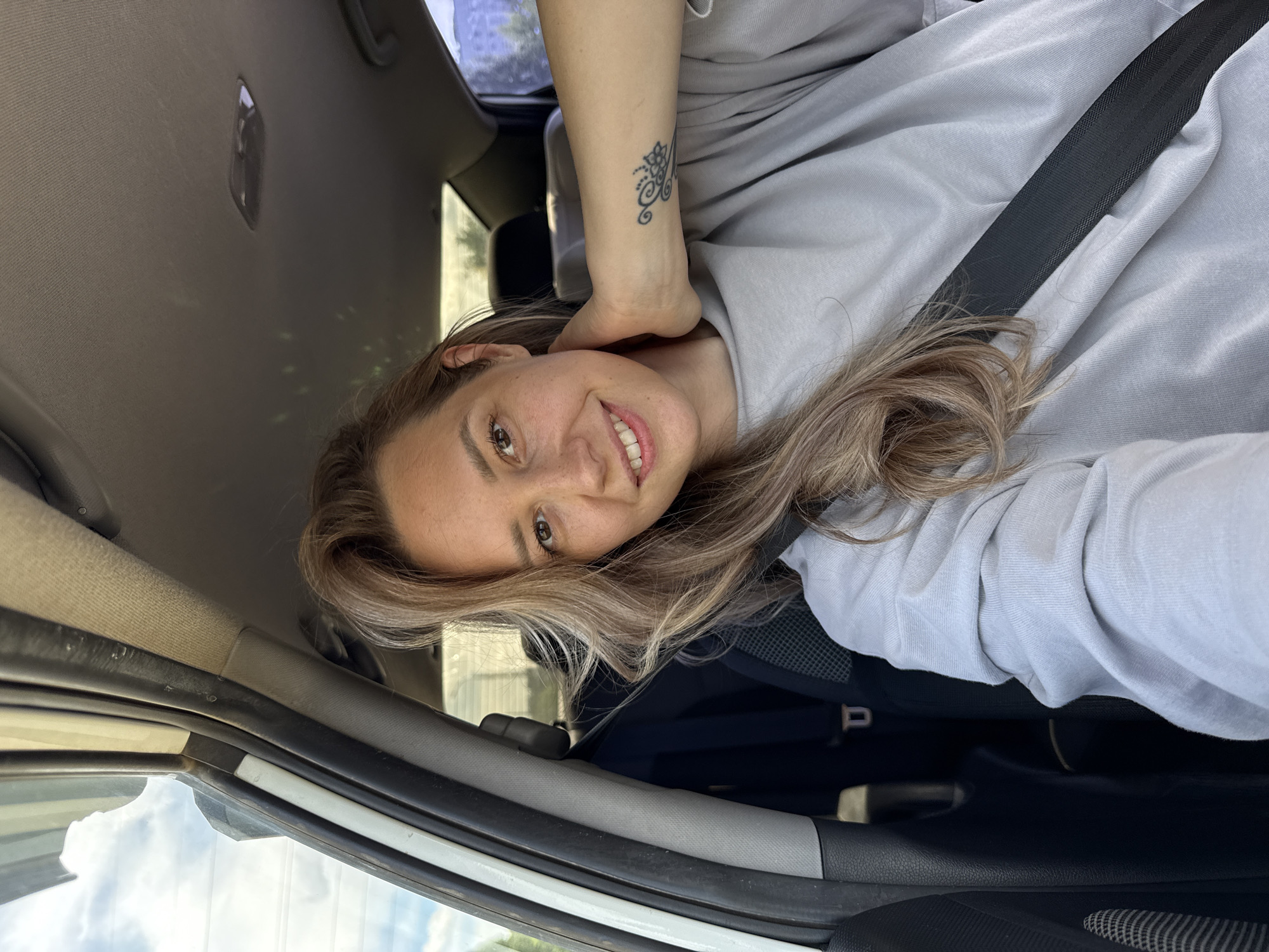 A woman with blonde hair smiles as she sits in the driver's seat of a car in Chaikovsky, Russia. She is wearing a light gray sweatshirt and has a black seatbelt fastened across her chest. A tattoo with a floral design is visible on her left forearm. The interior of the car, including the dashboard and headliner, is visible. The car appears to be parked, with daylight and a partly cloudy sky visible through the windows.