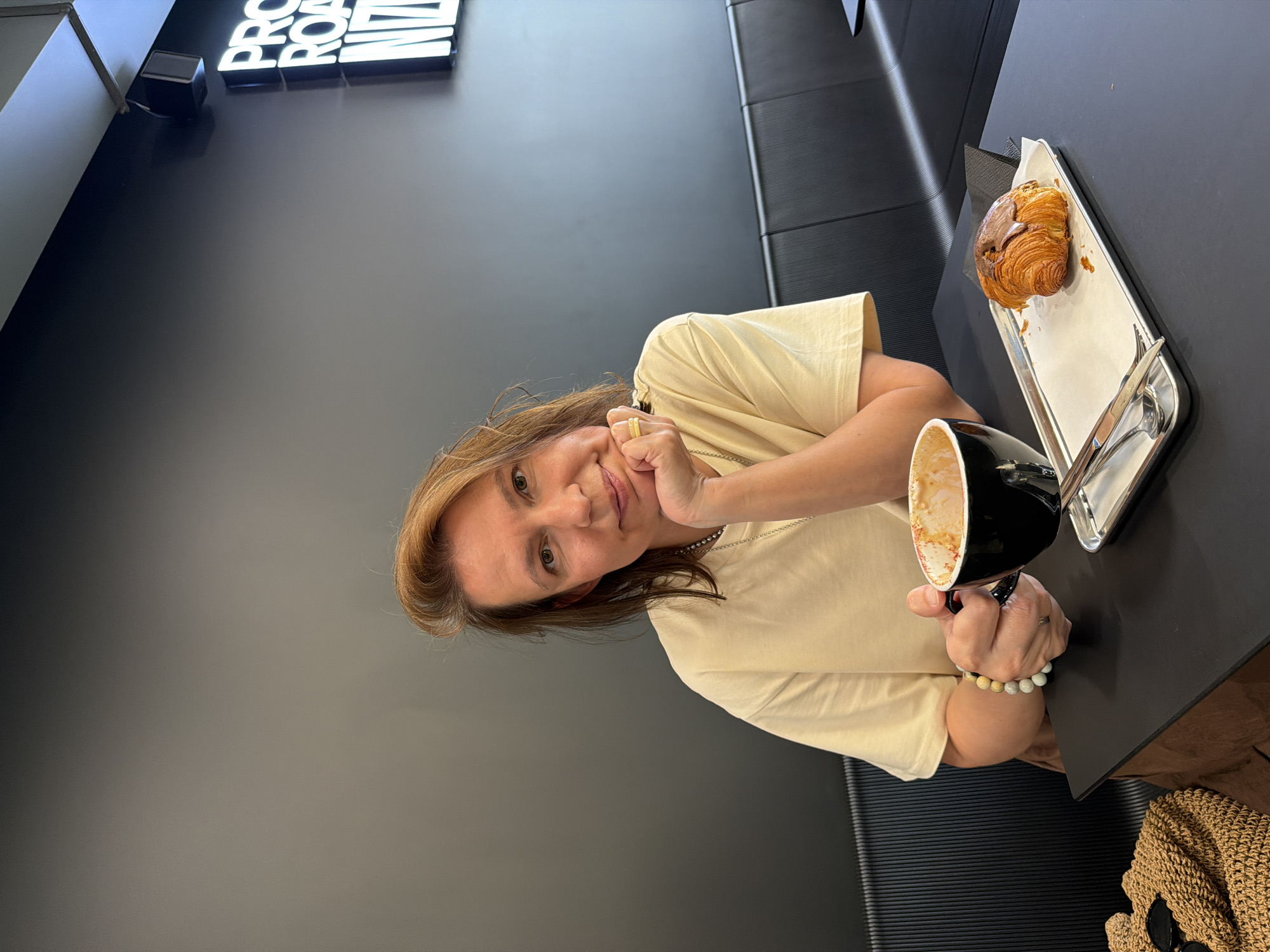 A medium shot shows a woman sitting at a table in a cafe in Izhevsk, Russia. She has light brown hair and is wearing a light yellow t-shirt and a brown skirt. Her left hand rests on her chin, and her right hand holds a black cup of coffee. A chocolate croissant is on a silver tray in front of her, next to a fork and knife. The background is a dark grey wall with a neon sign that reads "PRO ROAST NIZHNIY" in white letters. The woman appears to be looking at the camera and smiling.