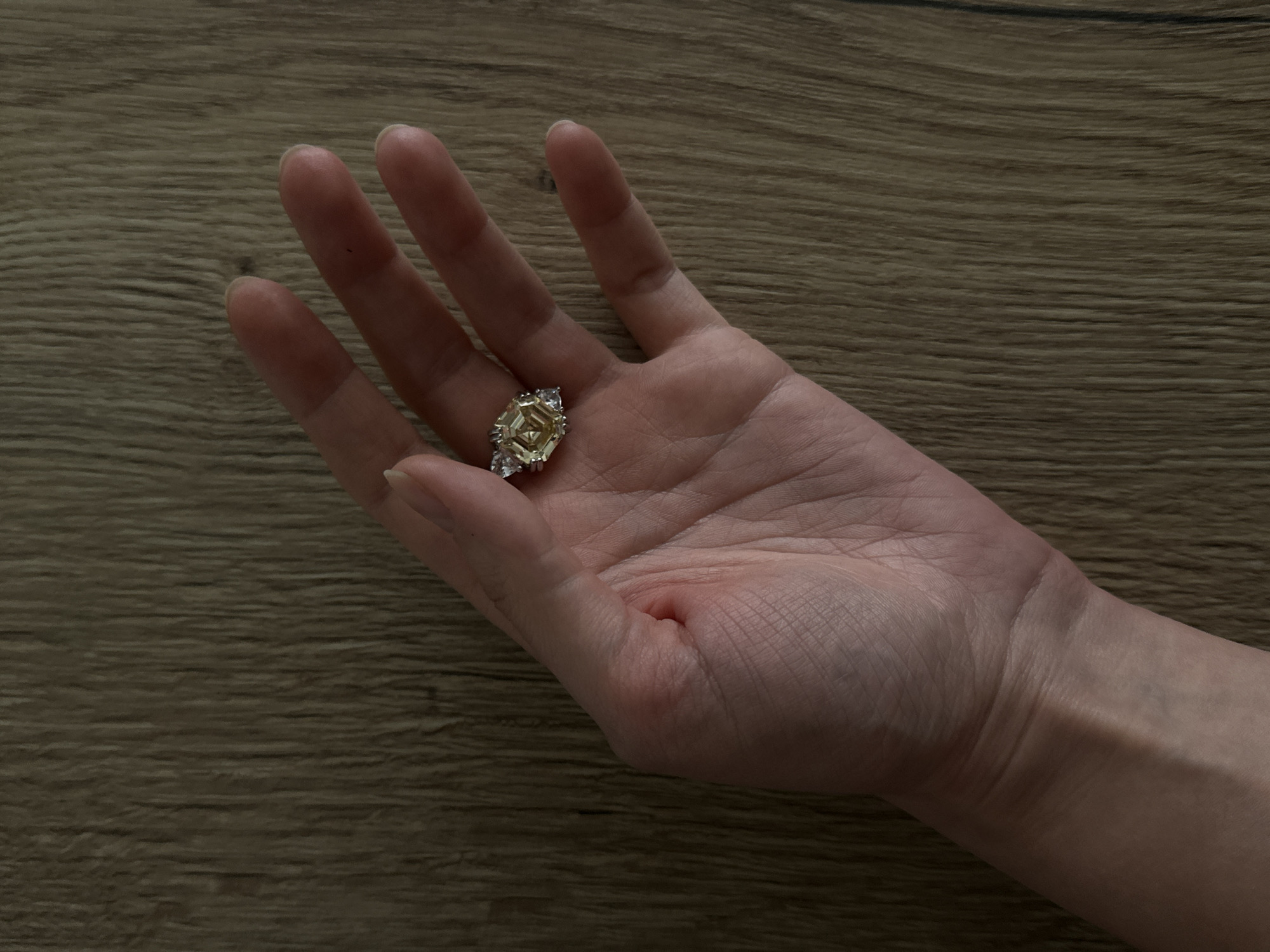 The image displays a close-up, high-angle shot of a human hand, held open with the palm facing upwards. The hand appears to be that of an adult, with visible skin texture and lines on the palm. Resting centrally in the palm is an elaborate ring. The ring features a large, multifaceted central stone with a distinct yellow or golden hue, possibly an emerald-cut or radiant-cut diamond. This central stone is accompanied by smaller, clear or white brilliant-cut stones set into the metallic band on either side, adding sparkle to the piece.
The background is a wooden surface, likely a table or floor, exhibiting a light to medium brown color with a pronounced horizontal wood grain pattern. The lighting is soft and even, illuminating the hand and the ring without harsh shadows, suggesting an indoor setting. There is no specific activity taking place other than the presentation or holding of the ring. No emotions are discernible from the hand's posture. No text is visible in the media. This scene is set in Moscow, Russia.