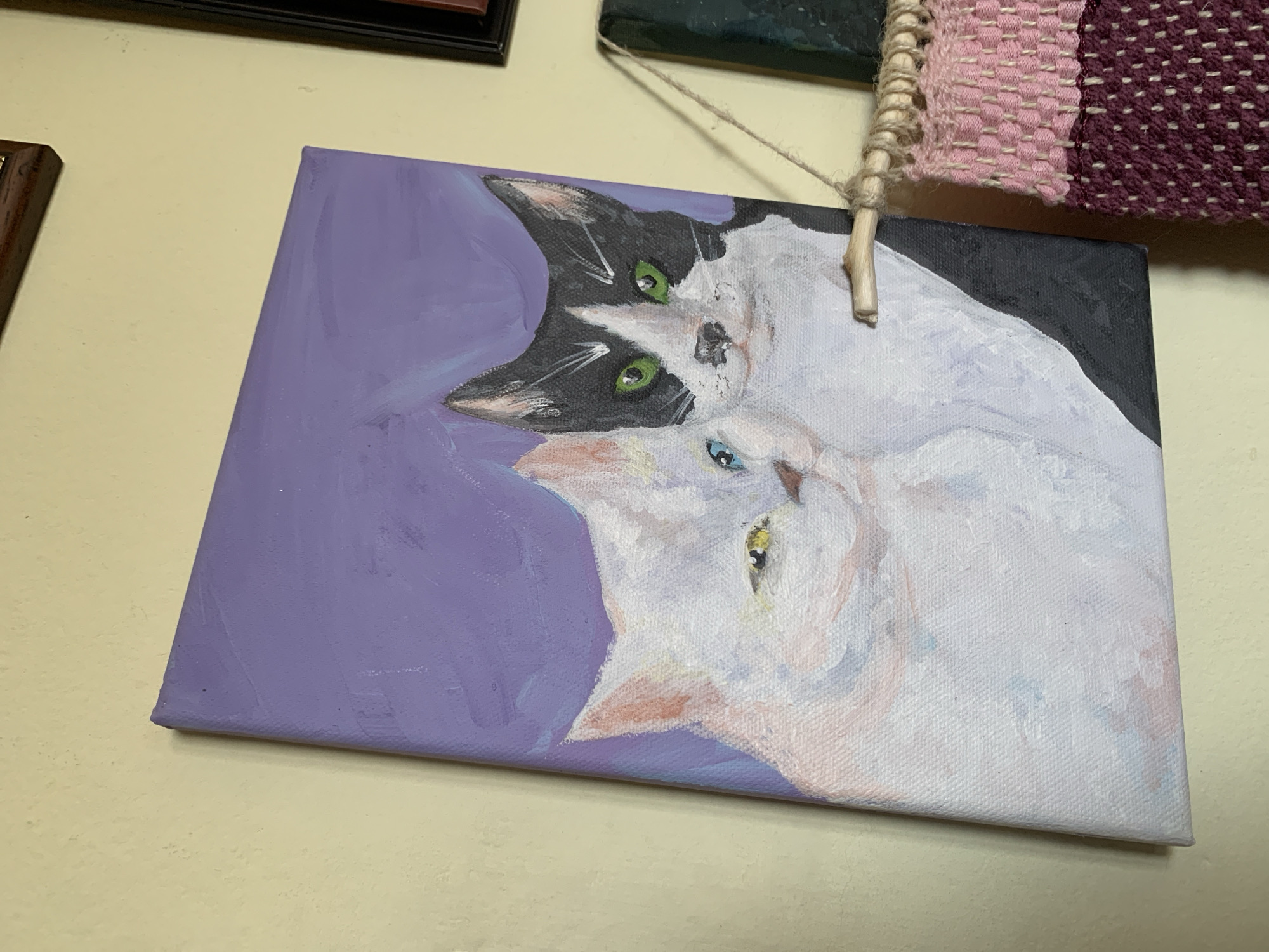 A painting of two cats hangs on a light yellow wall. The painting is a close-up of a black and white cat above a white cat, against a lavender background. A wooden stick with twine is attached to the top of the painting. The painting is hanging alongside other framed objects, including a woven piece with pink and purple tones. The location is unknown and no evidence suggests the city of Sofia, Bulgaria.