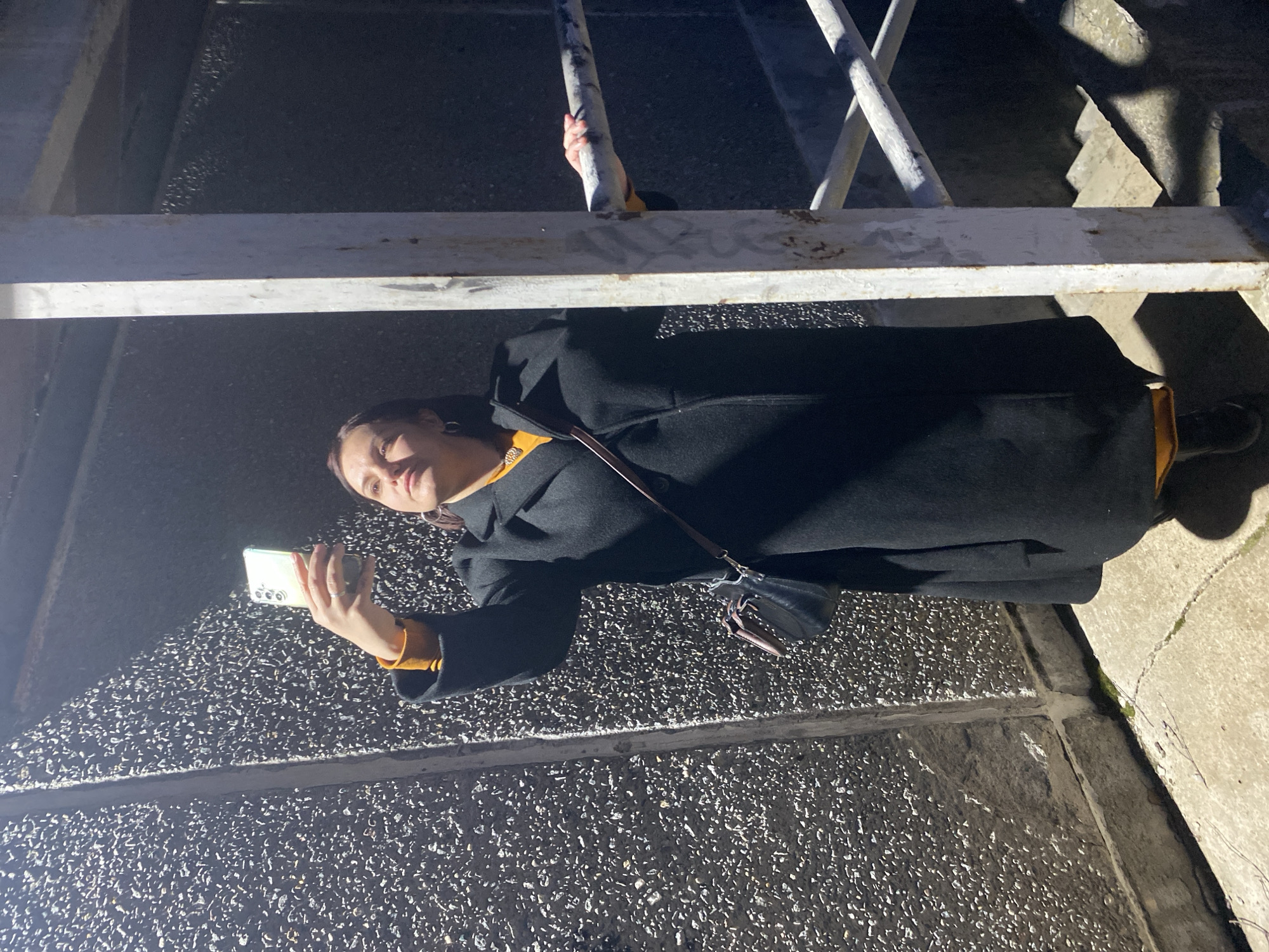 A woman stands on a sidewalk in Марковский, Russia, looking at her cell phone. She wears a black coat with a mustard-colored shirt underneath, dark shoes, and a crossbody bag. Above her is a metal structure of white bars. The sidewalk is made of textured concrete. The scene appears to be at night or in low light.