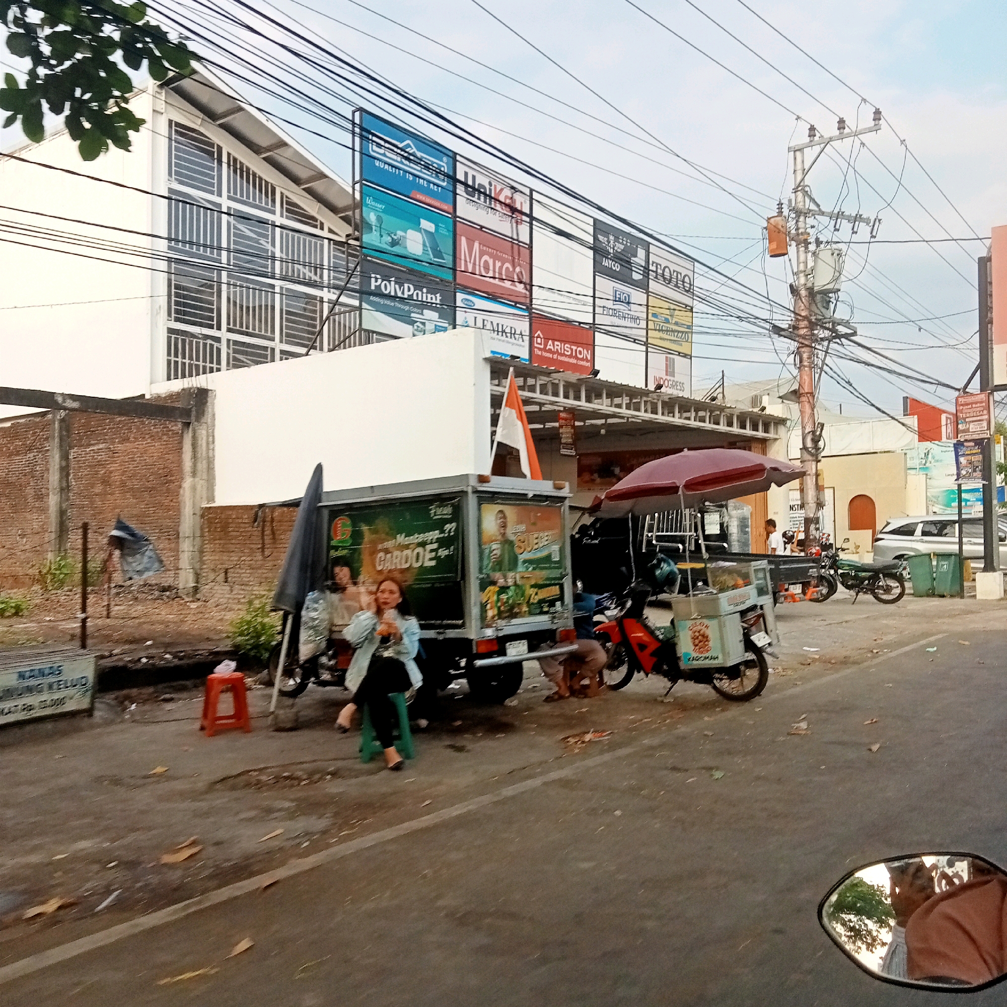 This image depicts a street scene in Karanganyar, Indonesia, likely during daylight hours with overcast skies. In the foreground, a paved road is visible with some debris and fallen leaves scattered on it. To the left, a white sign with blue lettering reads "NANAS DURIAN KELUD" and some Indonesian text. A small red stool is placed on the roadside.
The central focus of the image is a small truck with a white cargo box and colorful advertisements on its sides. The advertisements feature images of food and people. An Indonesian flag, orange and white, is attached to the top of the truck. Next to the truck, there is a food cart with a large red and white umbrella providing shade. A woman is seated on a green stool, holding a drink and looking towards the right. Another person, partially obscured, is bent down near the truck.
Behind the truck and food cart, there is a building with a white facade and a distinct architectural style, featuring a sloped roof with many windows. Above this building, a series of large billboards are mounted, displaying various brand names and logos, including "Unikall," "Marco," "TOTO," "Jayco," "Fiorentino," "Vicenzzo," "Lemkra," and "Ariston."
To the right of the truck, a red and black motorcycle is parked, and further down the street, a green motorcycle is visible. A white car is parked on the right side of the road, next to two green trash bins. A utility pole with numerous electrical wires and transformers stands prominently on the right side of the image. In the distance, more buildings and signs can be seen. A motorcycle's side mirror in the bottom right corner reflects a person's face. The overall impression is that of a busy commercial area with a mix of vehicles, businesses, and street vendors.
