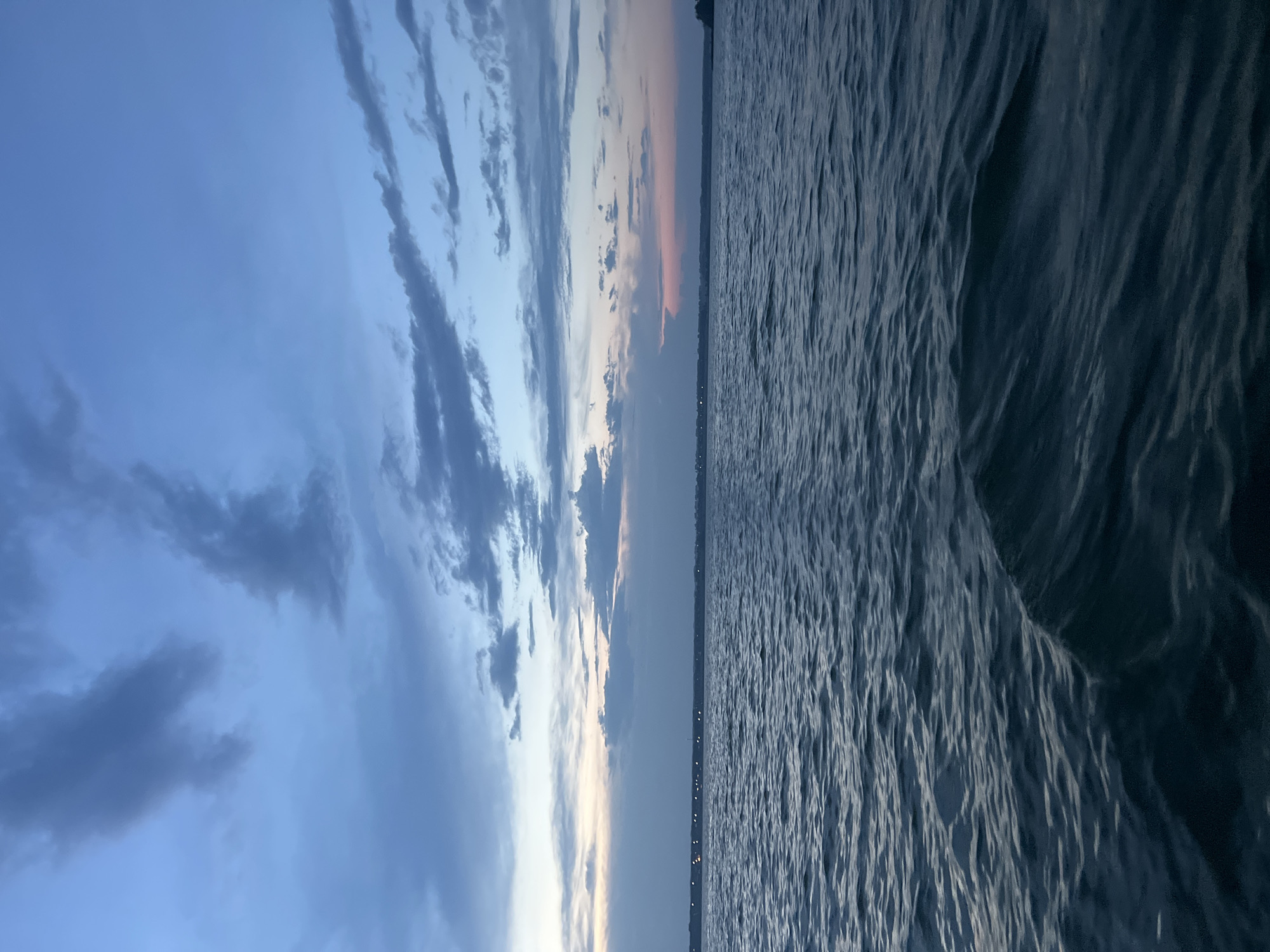 The image depicts a body of water under a twilight sky. The water is dark and appears to be choppy, with visible ripples and a small wave in the foreground. In the distance, a faint line of lights suggests a shoreline or a settlement. The sky is a deep blue with scattered clouds that are tinged with pink and gray hues, indicating the sun has recently set or is about to rise. The overall atmosphere is serene and tranquil. The location is not discernible from the image itself, and any mention of Novopichugovo, Russia, is not supported by visual evidence.