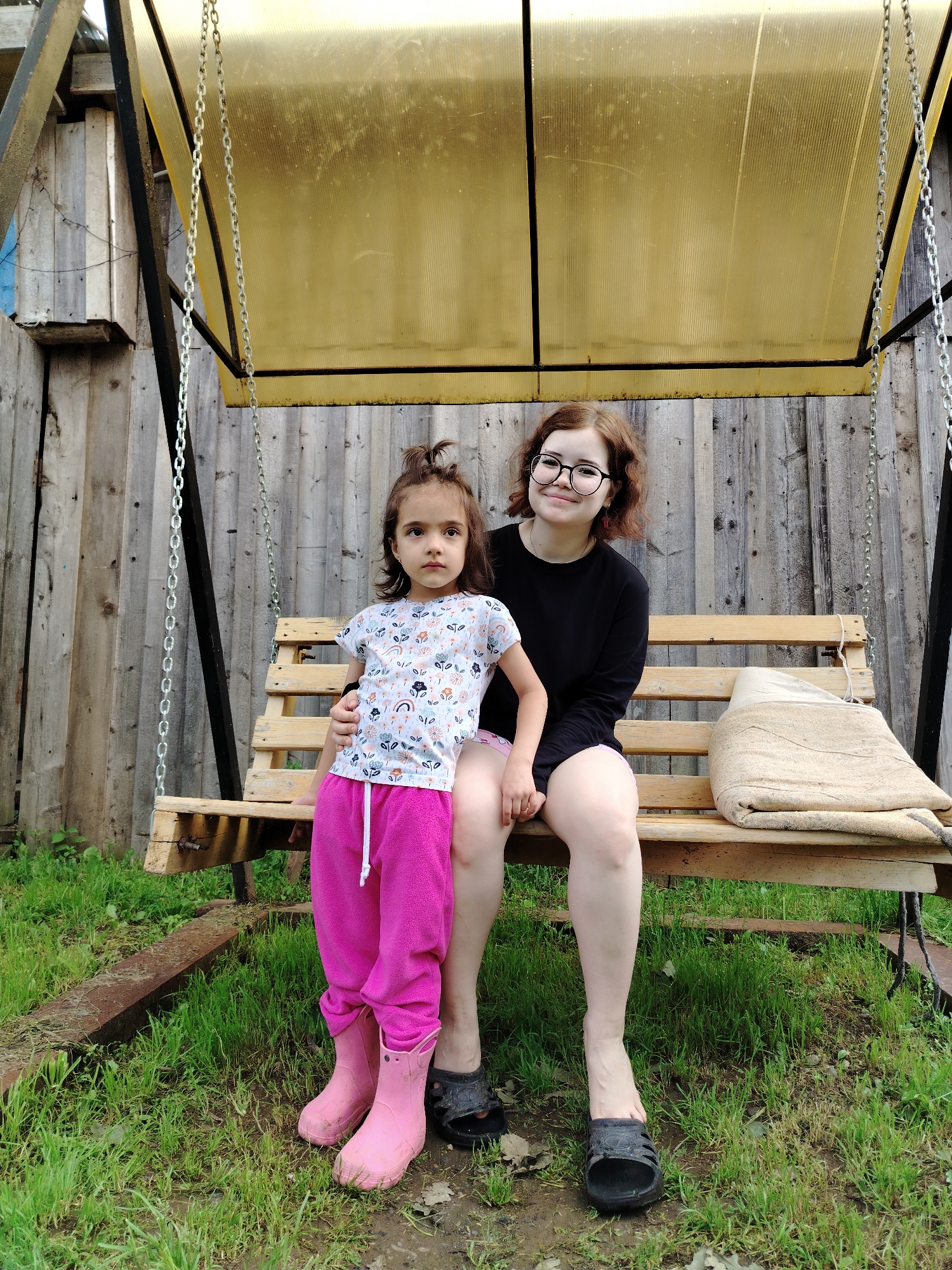 Two females, an adult and a child, are seated on a wooden swing. The adult, with reddish-brown curly hair and glasses, wears a black long-sleeved shirt and is positioned on the right. The child, with dark brown hair tied in a messy bun, wears a white t-shirt with a colorful pattern and bright pink pants, and stands on the left, leaning slightly against the adult. Both are sitting on the wooden slats of the swing, which is suspended by metal chains from a frame with a yellow, ribbed plastic roof. A beige, folded blanket or cushion rests on the right side of the swing seat. The background consists of a weathered wooden fence and lush green grass. The setting appears to be a yard or backyard, possibly in the city of Gorzhan, Russia, given the style of the fence and the overall rustic environment.