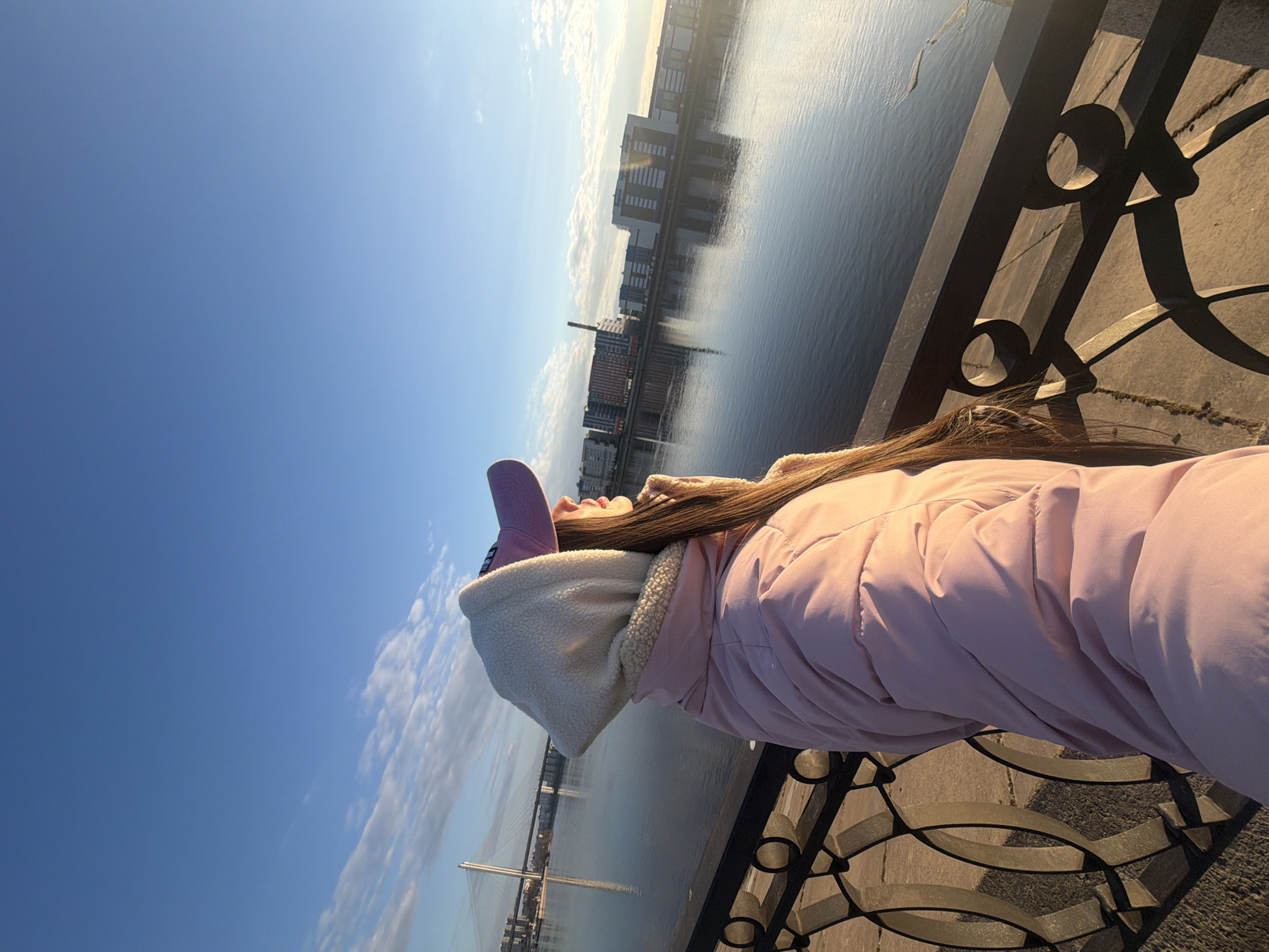 A woman in a pink puffer jacket and a lavender hat stands on a bridge in Saint Petersburg, Russia, looking up at the sky. She is leaning against a black metal railing with a decorative design. In the background, a body of water reflects the buildings across the river. A cable-stayed bridge is visible in the distance. The sky is blue with some clouds.