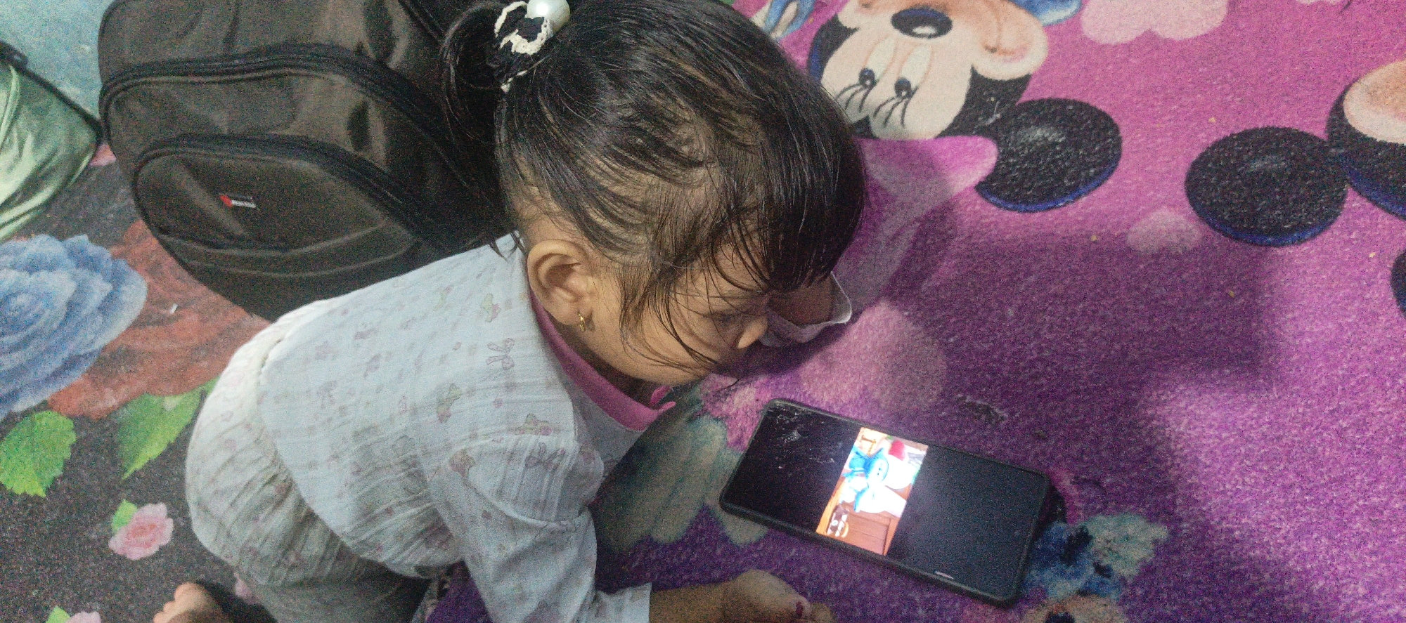 A toddler wearing a light blue pajama with a floral pattern, crouches on a pink blanket featuring a cartoon character. The child is looking intently at a smartphone that displays a cartoon image. A dark backpack is situated behind the child, and the overall scene suggests a domestic setting, possibly a living room or bedroom, within the City of Pematang Siantar, Indonesia.