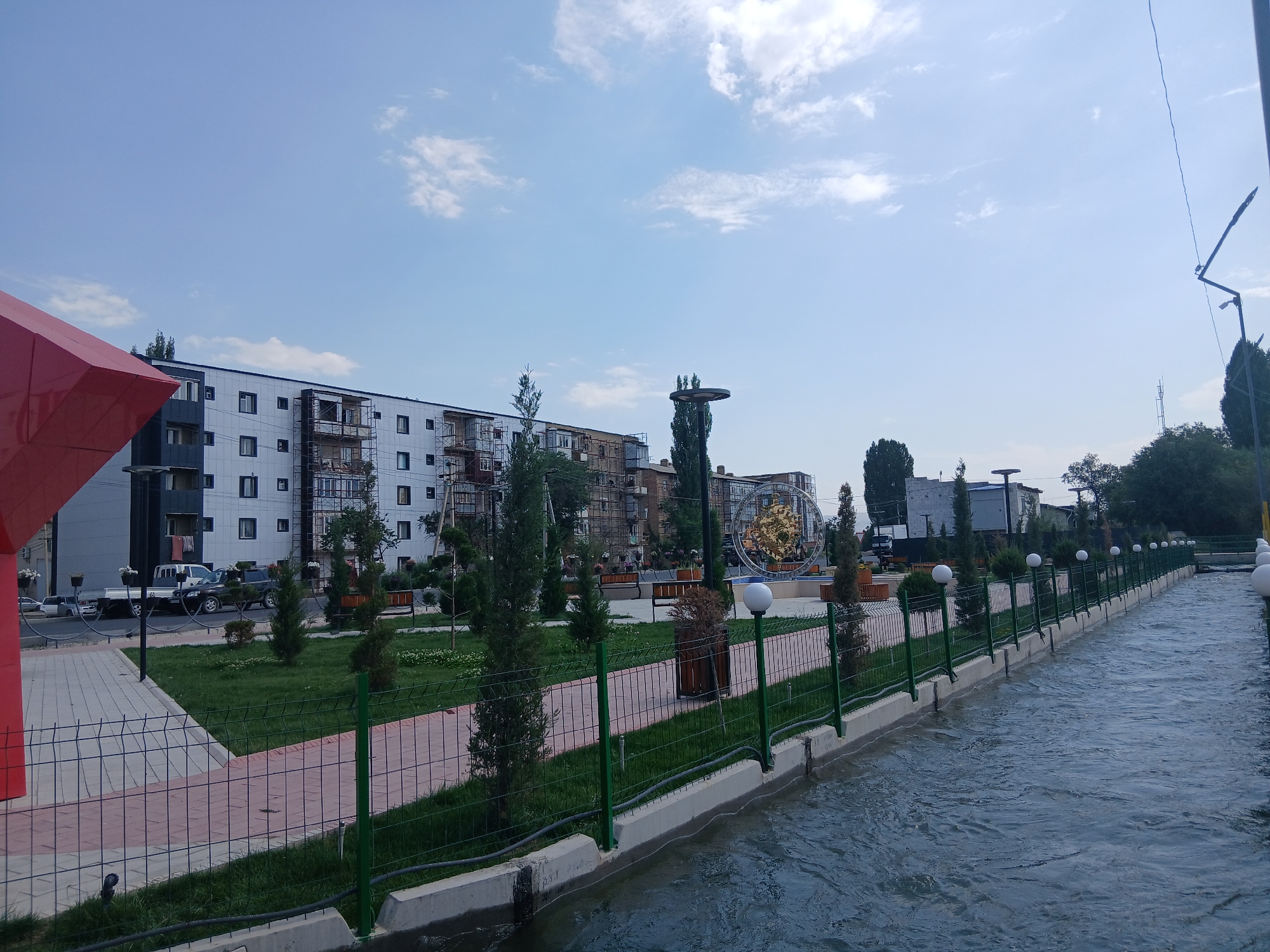 The image depicts a canal running through an urban area in Talas City, Kyrgyzstan. The canal is bordered by a concrete embankment and a green metal fence. A paved walkway runs alongside the canal, separated by a lawn and more fencing. In the background, a modern, multi-story building complex with white and dark gray facades is visible. The building appears to be under construction or renovation, with scaffolding on some sections. A large, circular golden sculpture or art installation stands in the middle ground. Several trees and lampposts are scattered throughout the scene. The sky is light blue with some scattered white clouds. A red structure, possibly a canopy or art piece, is partially visible in the foreground on the left. The water in the canal is moving, suggesting a current. There are no people visible in the image.