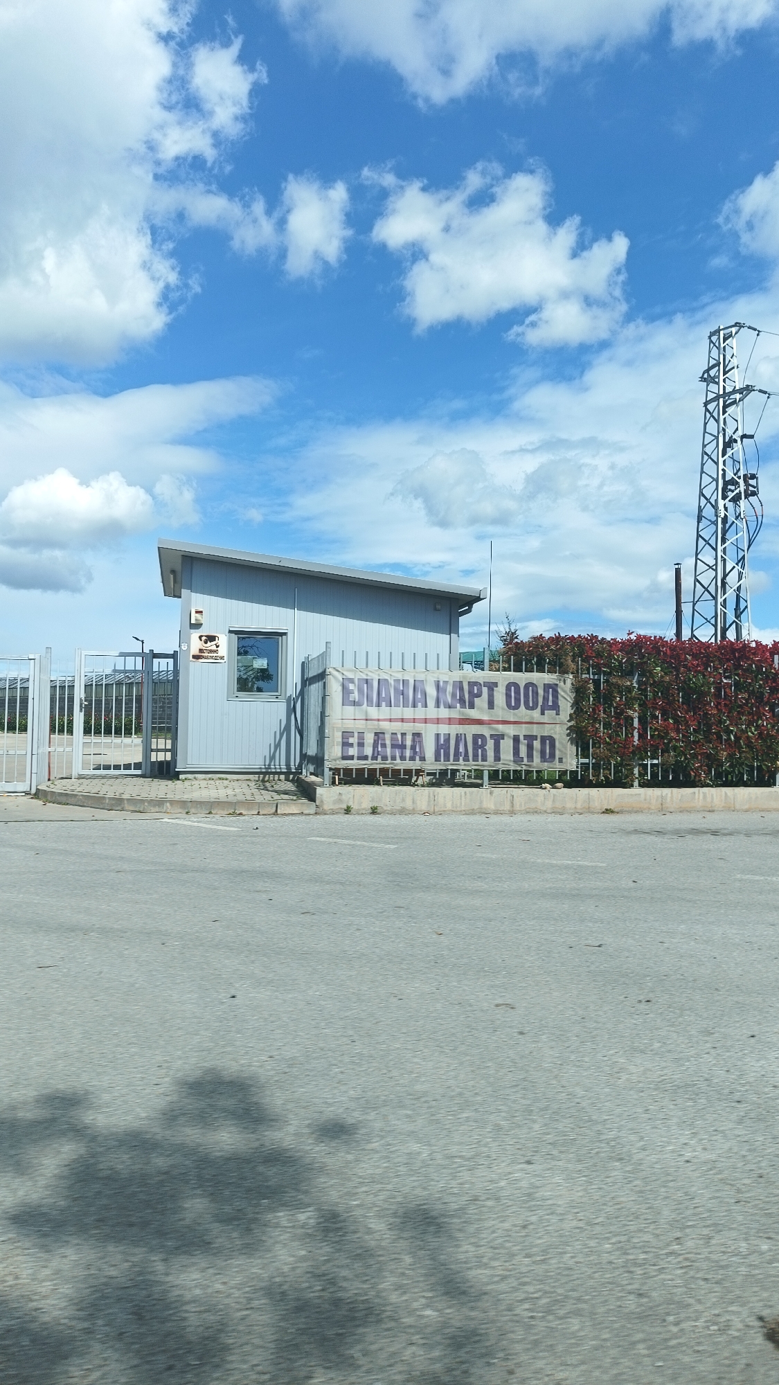 The image shows the entrance to ELANA HART LTD, located in Boliartsi, Bulgaria. A light blue guardhouse sits beside a metal fence displaying a sign with the company name in both Cyrillic and Latin alphabets. To the right of the fence is a hedge of red-leaved plants. An electrical tower is visible in the background. The foreground is a paved road surface with shadows cast onto it. The sky is blue with scattered white clouds. There are no people visible in the image.