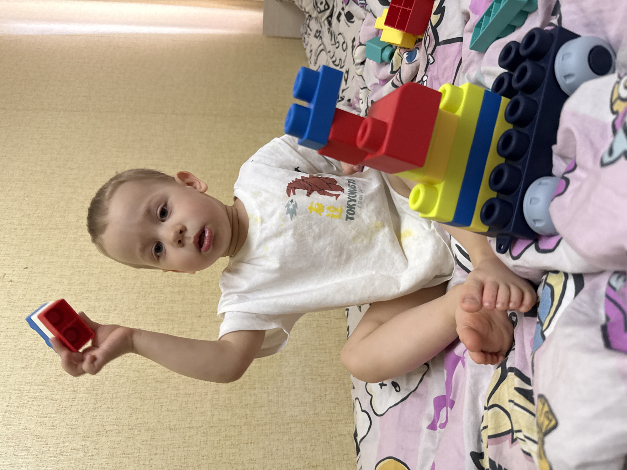 A young child is depicted lying on their side on a bed in a domestic setting, likely in Rusanovo, Russia. The child has light hair and fair skin, and is wearing a white t-shirt featuring a brown graphic of a creature and text that includes "TOKYO" and "MONSTER".
The child is actively playing with colorful building blocks. In their left hand, they hold a small red and blue block. Next to them on the bed, a larger construction of blocks (red, yellow, blue, and black) with wheels is visible, along with another turquoise block. The bed is covered with a vibrant, patterned duvet featuring cartoon-like characters, predominantly in pink, purple, and yellow tones. The background shows a light yellow, textured wallpaper.
The child is looking directly at the camera with a slightly open mouth, conveying a sense of curiosity and engagement. The scene appears to be taking place during the daytime, indoors.