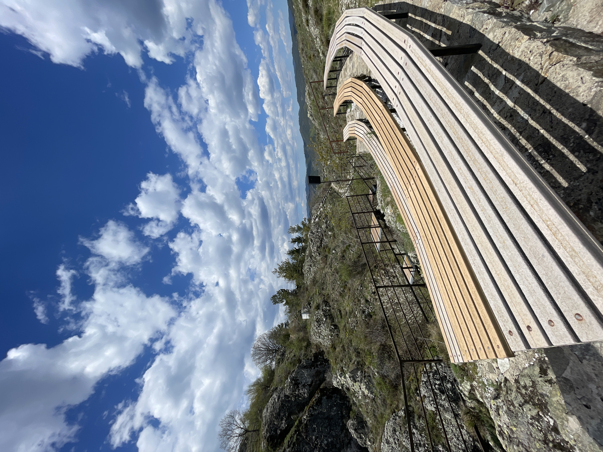The image shows a wooden bench and metal railing on a rocky cliff edge in Gradat, Bulgaria. The benches are made of light-colored wooden slats. Below the bench, there is a metal safety railing and a steep, rocky terrain with sparse vegetation. In the distance, there are hints of more hills and trees. The sky is bright blue with puffy white clouds. The photograph is taken from a high angle, looking downward.